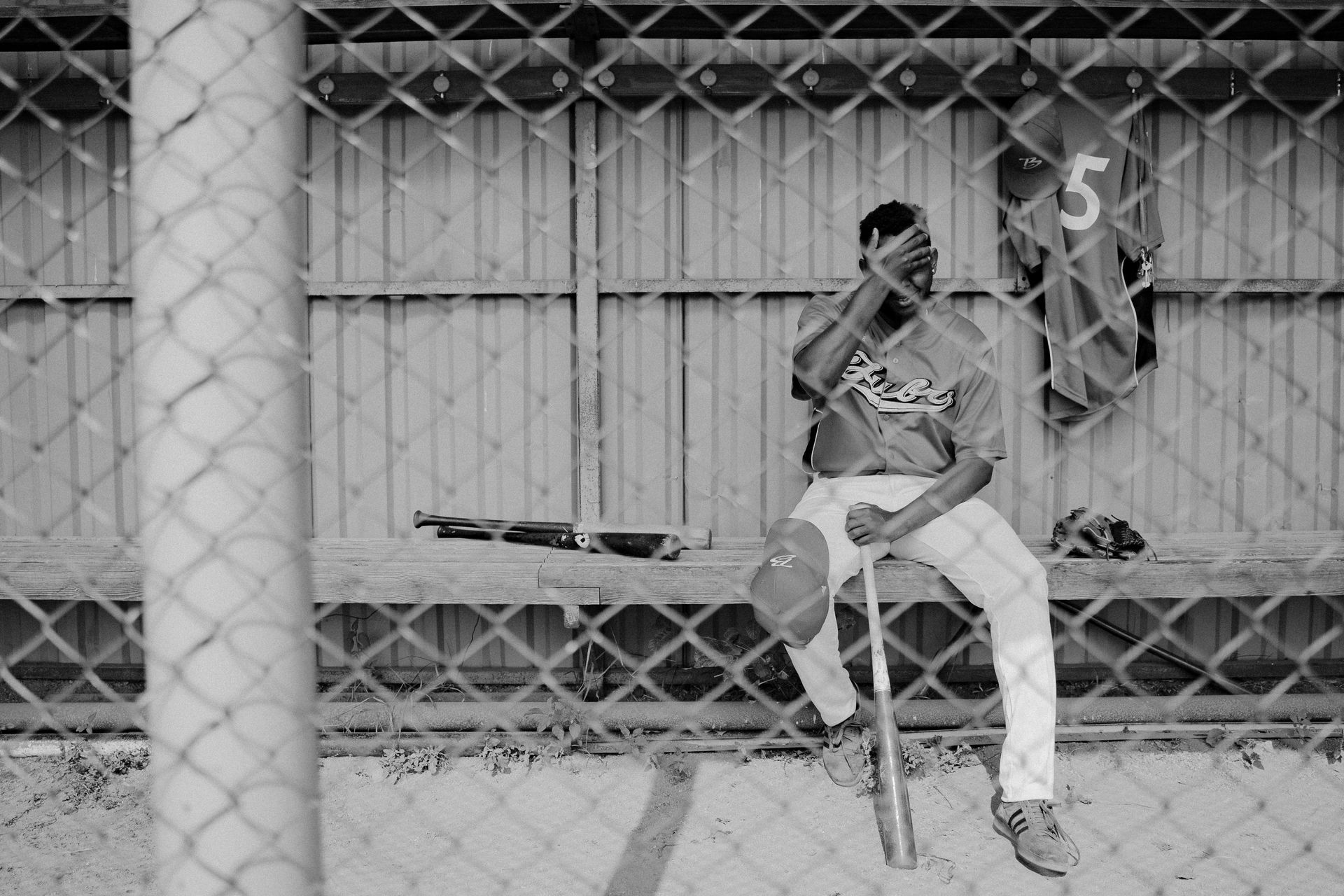 Baseball player sits on bench, holding bat, jersey hanging nearby behind a chain link fence.