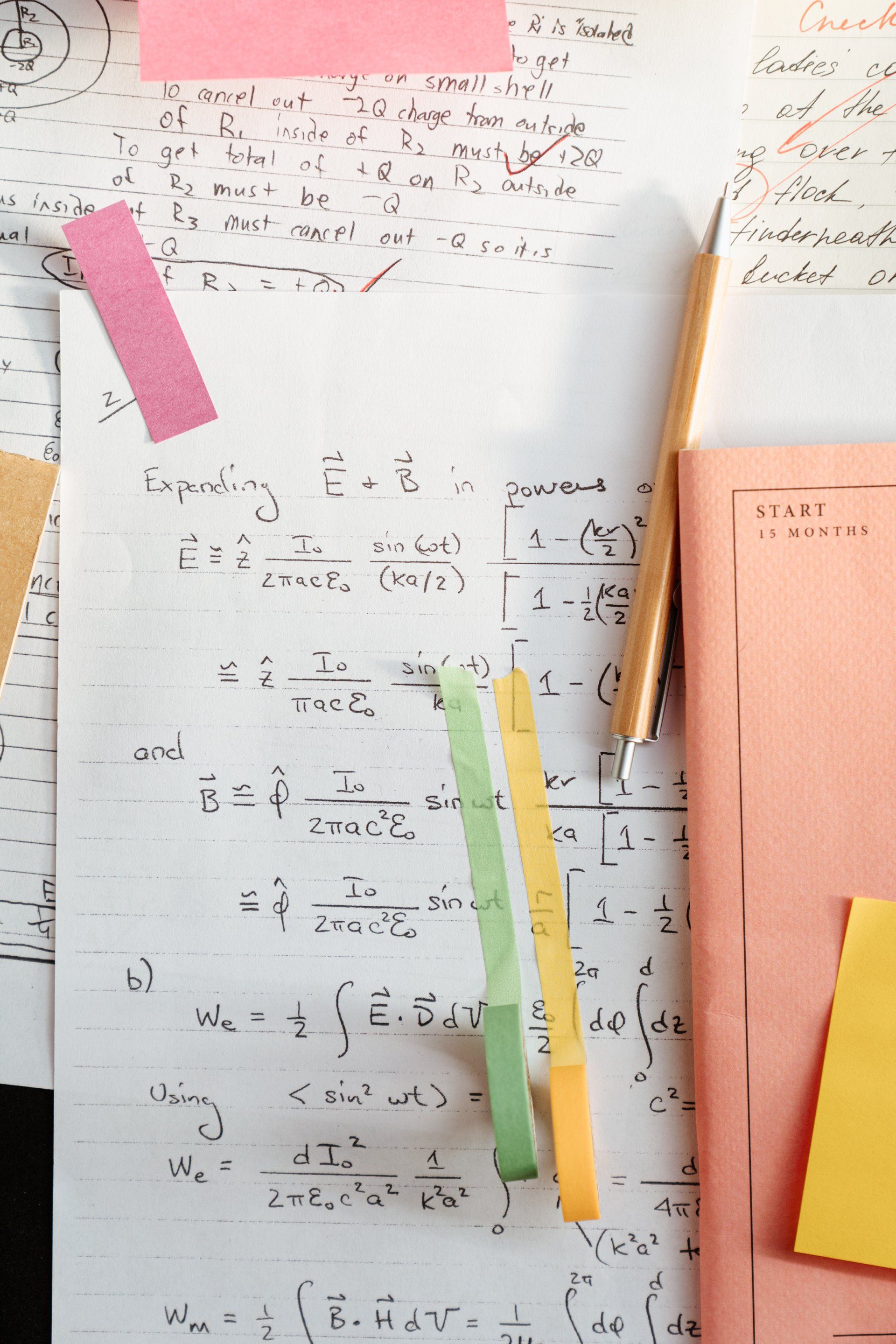 Close-up of handwritten math equations on paper with sticky notes, a pen, and a book.