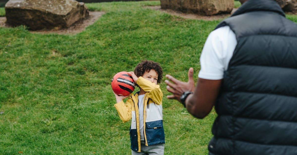 Child throwing a red ball to a person in a vest, on a grassy lawn with rocks.