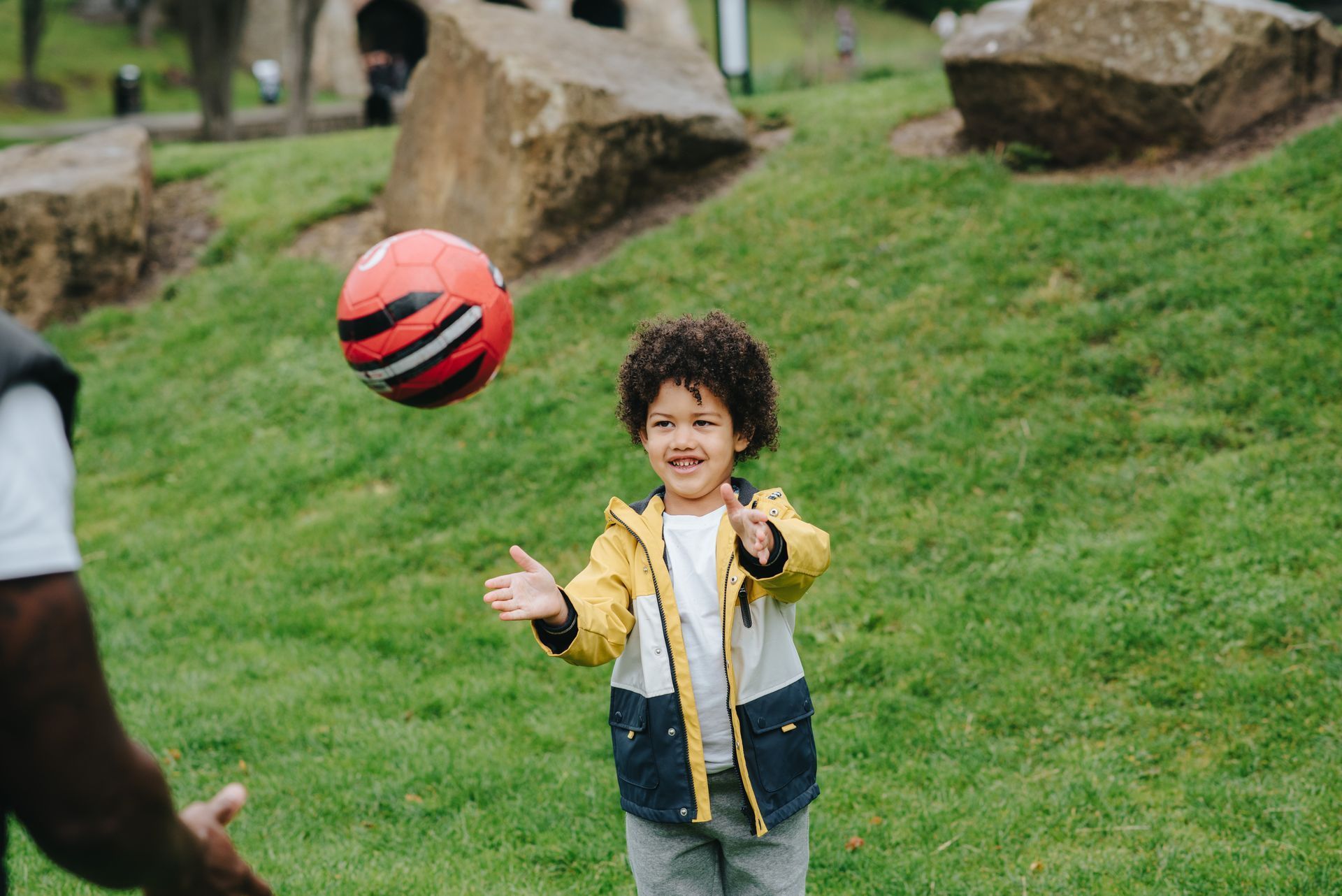 Child in yellow jacket smiling, catching a red and black ball in a grassy park.