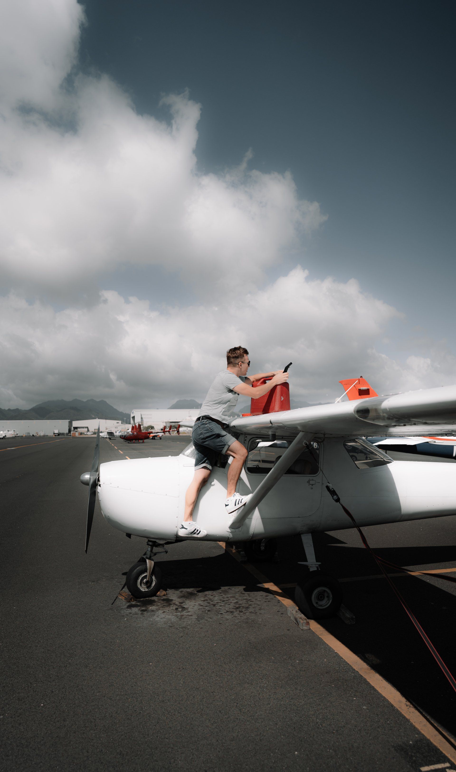 Person climbs onto the wing of a small airplane on a tarmac, holding a red object. Cloudy sky.