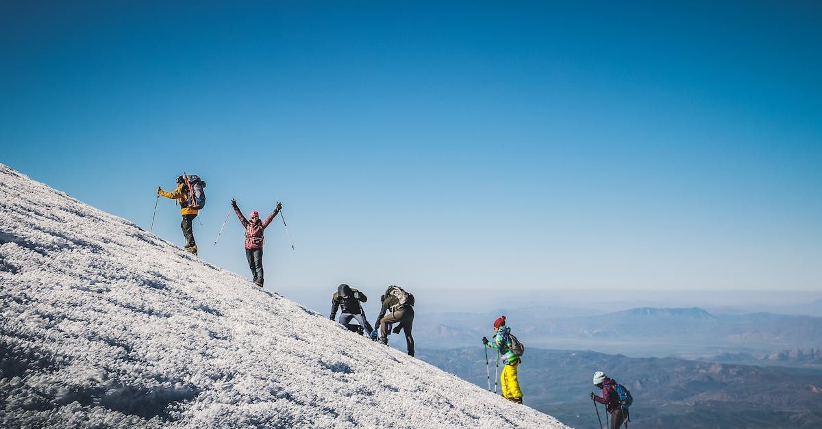 Hikers ascending snow-covered mountain under a clear, blue sky; one celebrates with arms raised.