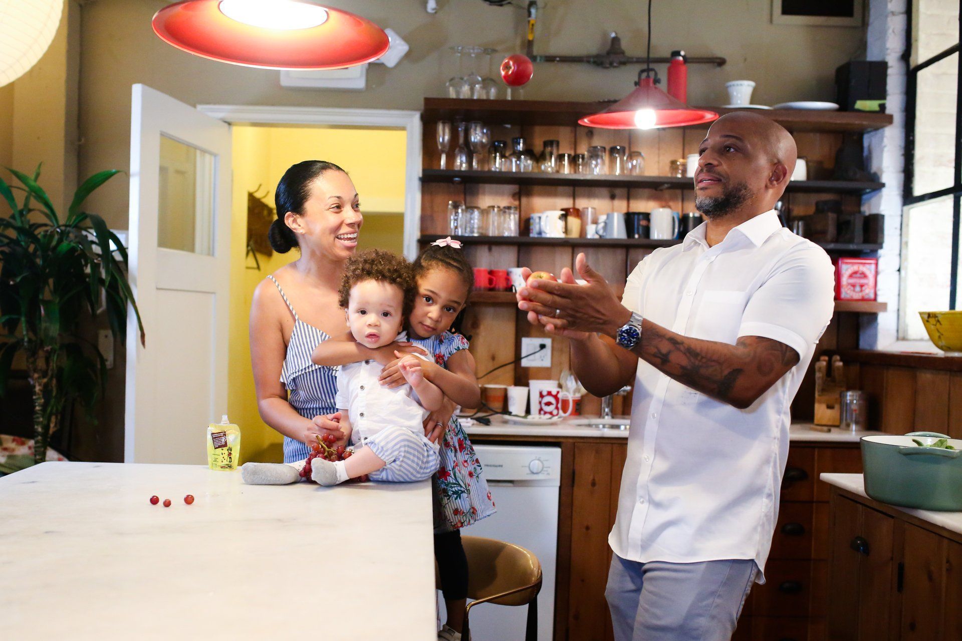 Family in a kitchen. Man clapping; woman holding two children, standing near a counter. Sunlight through a window.
