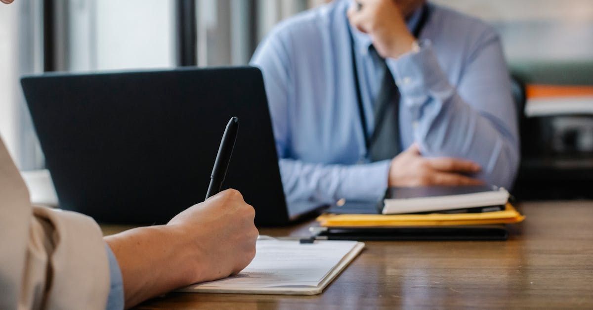 Person writing on paper at a table, facing another person with a laptop and documents; indoor office setting.