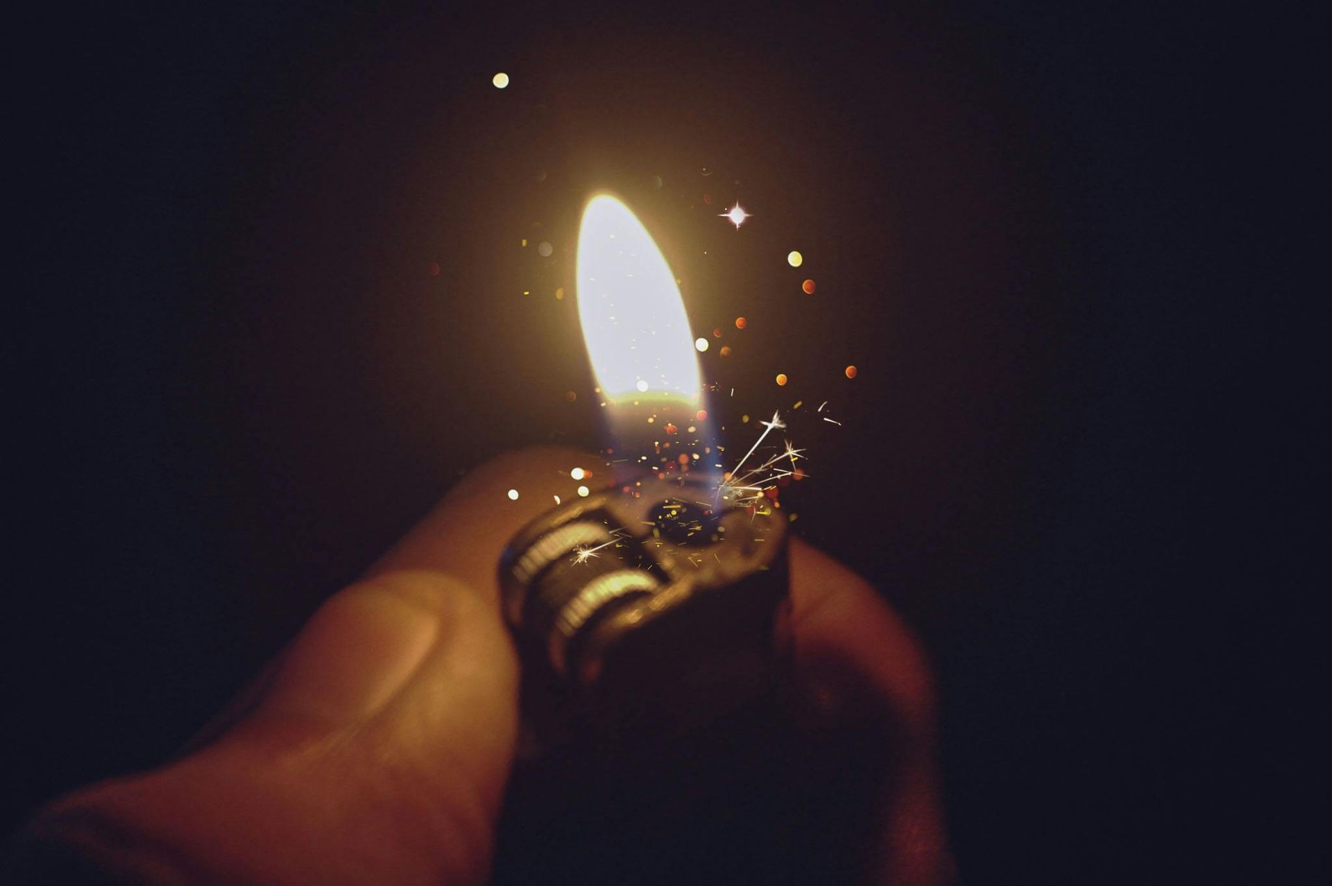 Hand holding a lit lighter in the dark, sparks flying from the flame.