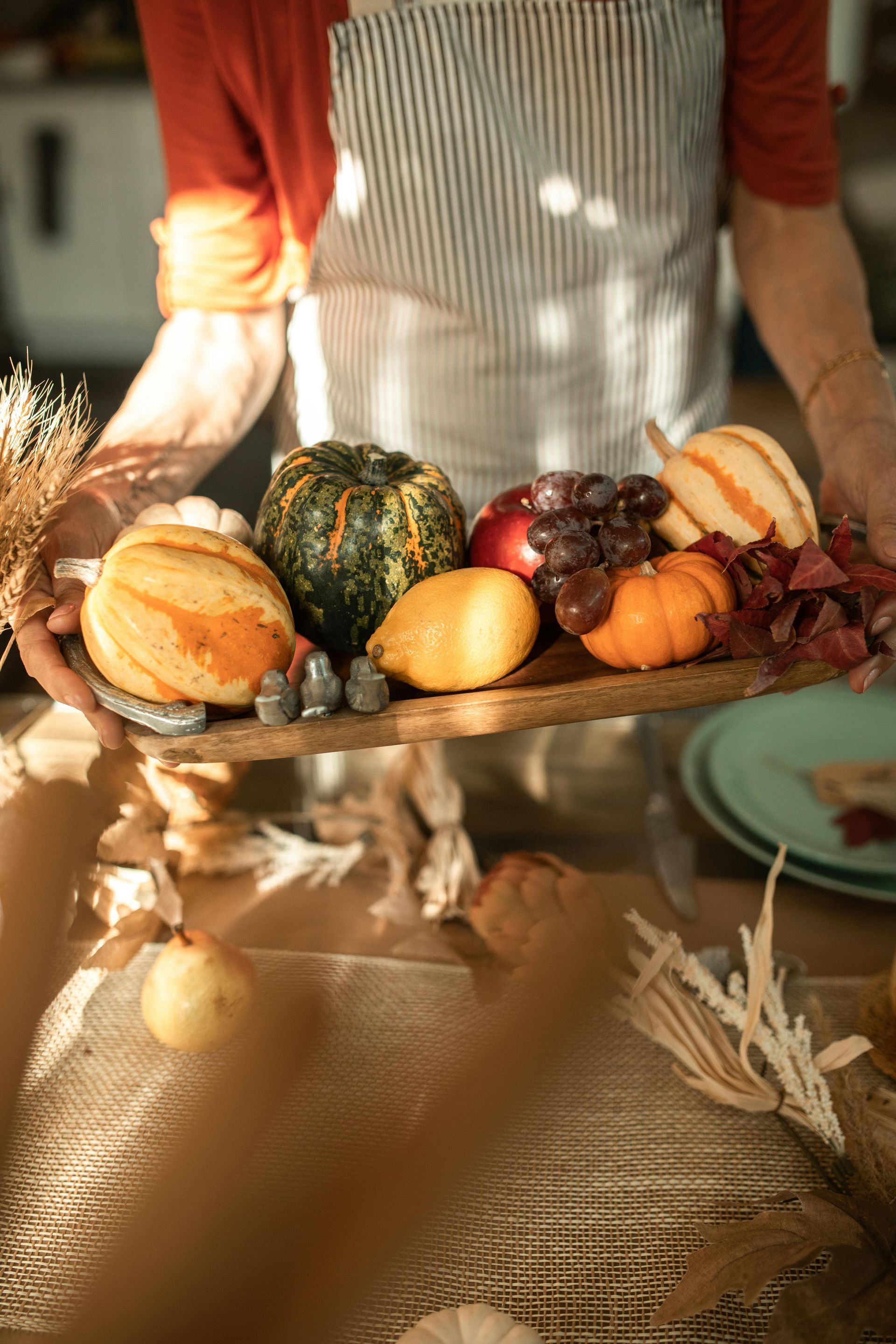 Person holding a wooden tray with gourds, apples, and grapes. Table setting.