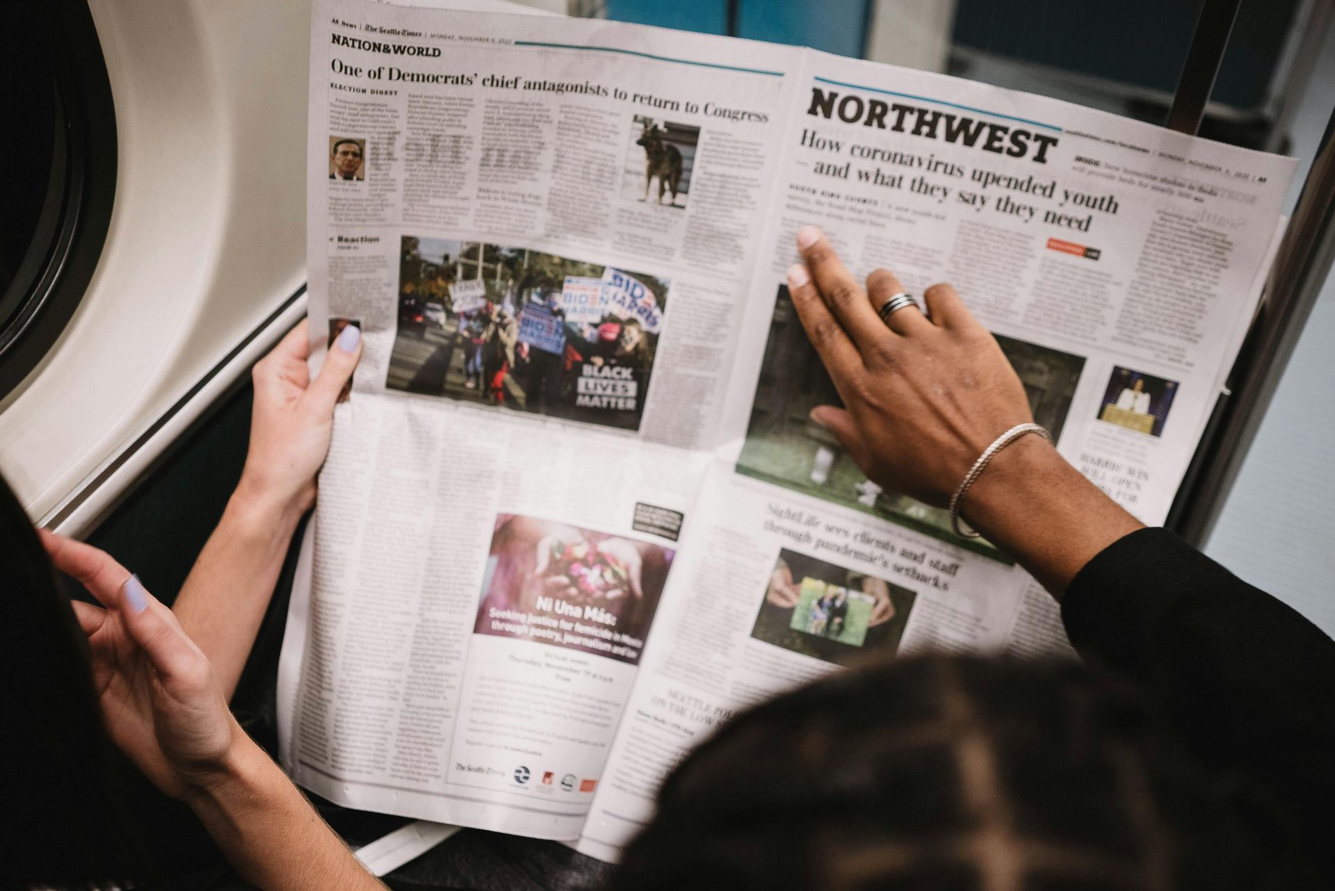 Person holding and reading a newspaper with headlines and photos; inside a public transit vehicle.