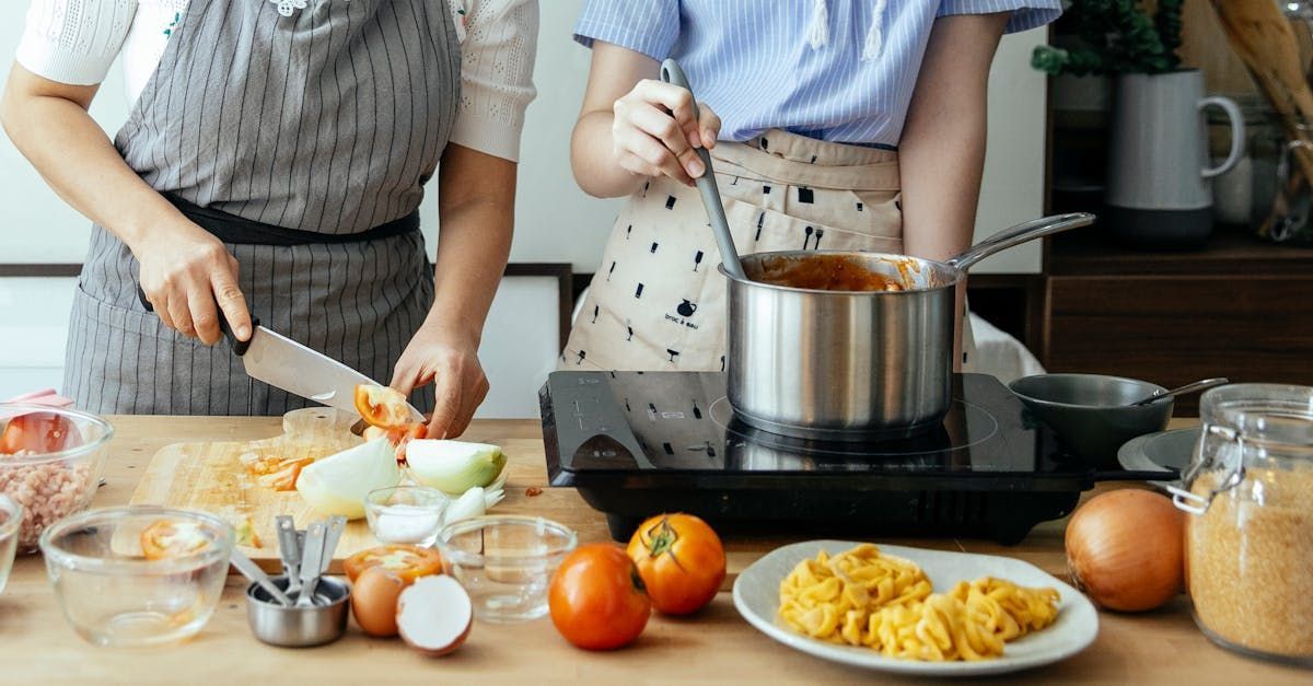 Two people cooking; one chopping vegetables, the other stirring sauce in a pot on a stovetop.