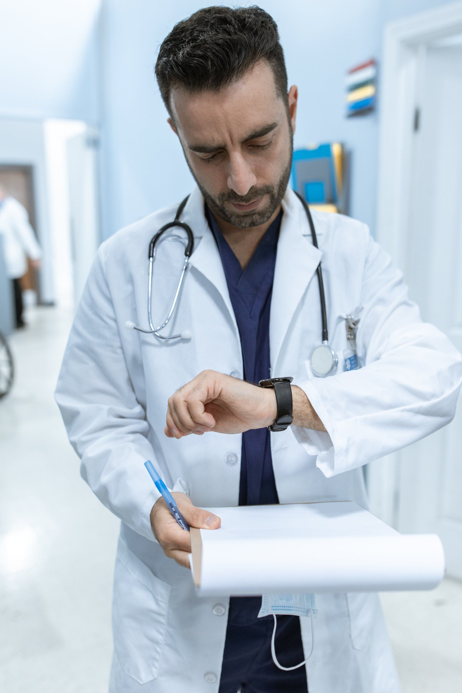 Doctor in a white coat looks at watch while holding a clipboard in a hospital hallway.