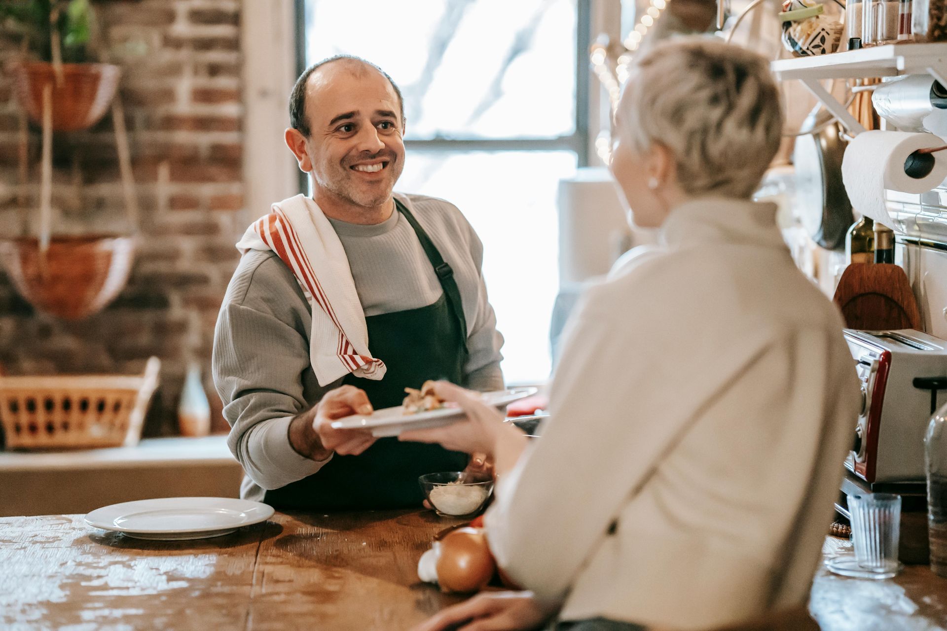Man in apron passing a plate of food to a person in a kitchen.