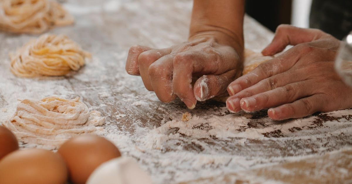 Hands kneading dough on a flour-dusted surface, with fresh pasta and eggs nearby.