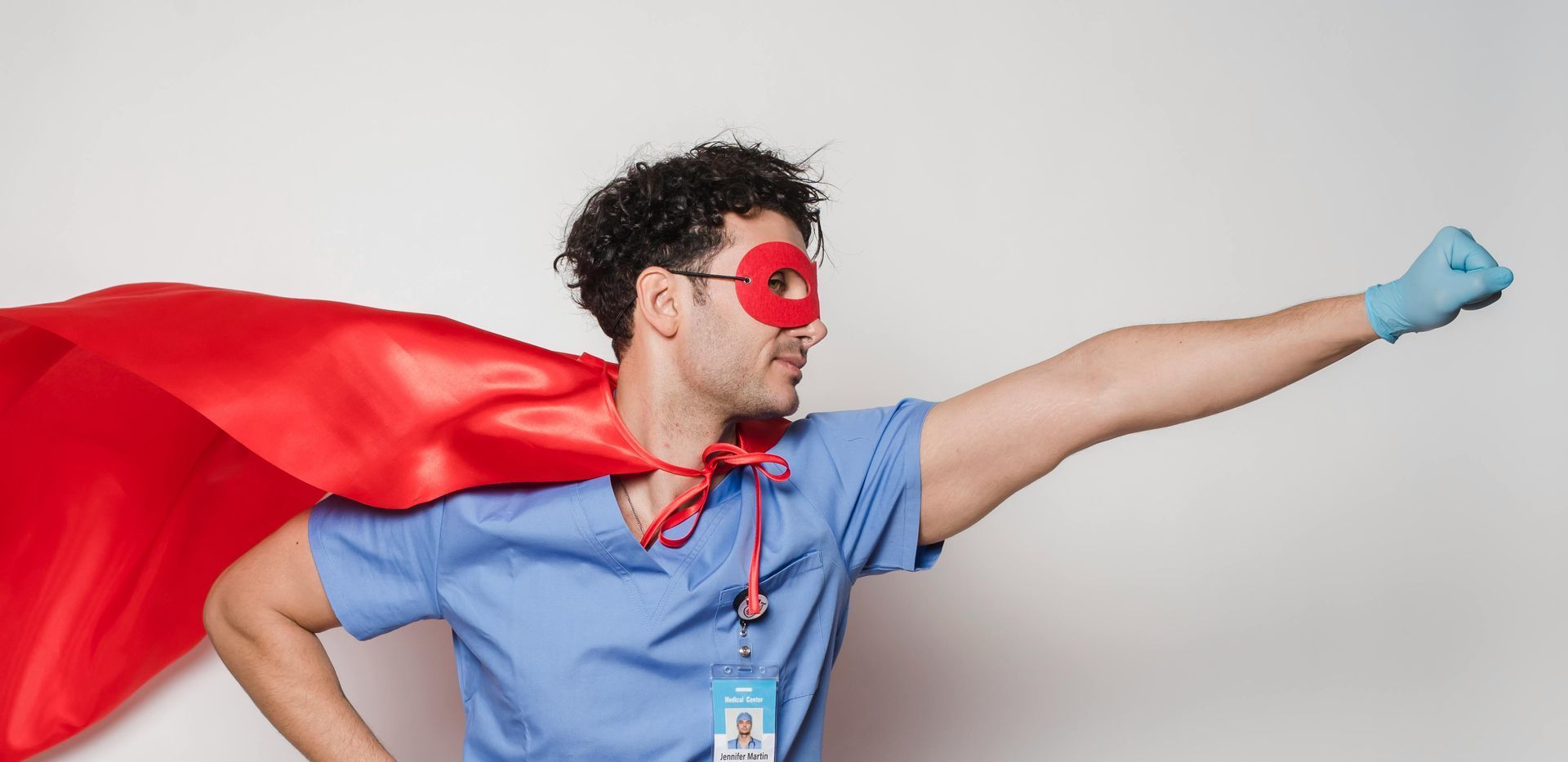 Healthcare worker in scrubs with a superhero mask and cape, arm outstretched in a flying pose.