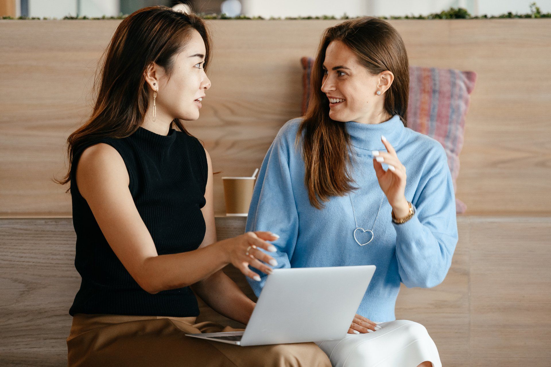 Two women seated, discussing at laptop. Woman in black points; woman in blue smiles, gesturing.