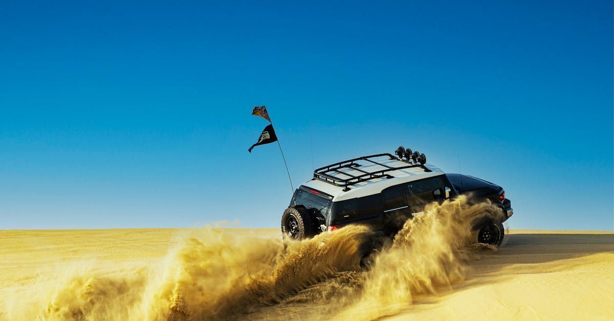 Black SUV driving across a sandy desert, kicking up sand, kite in the background, against a blue sky.
