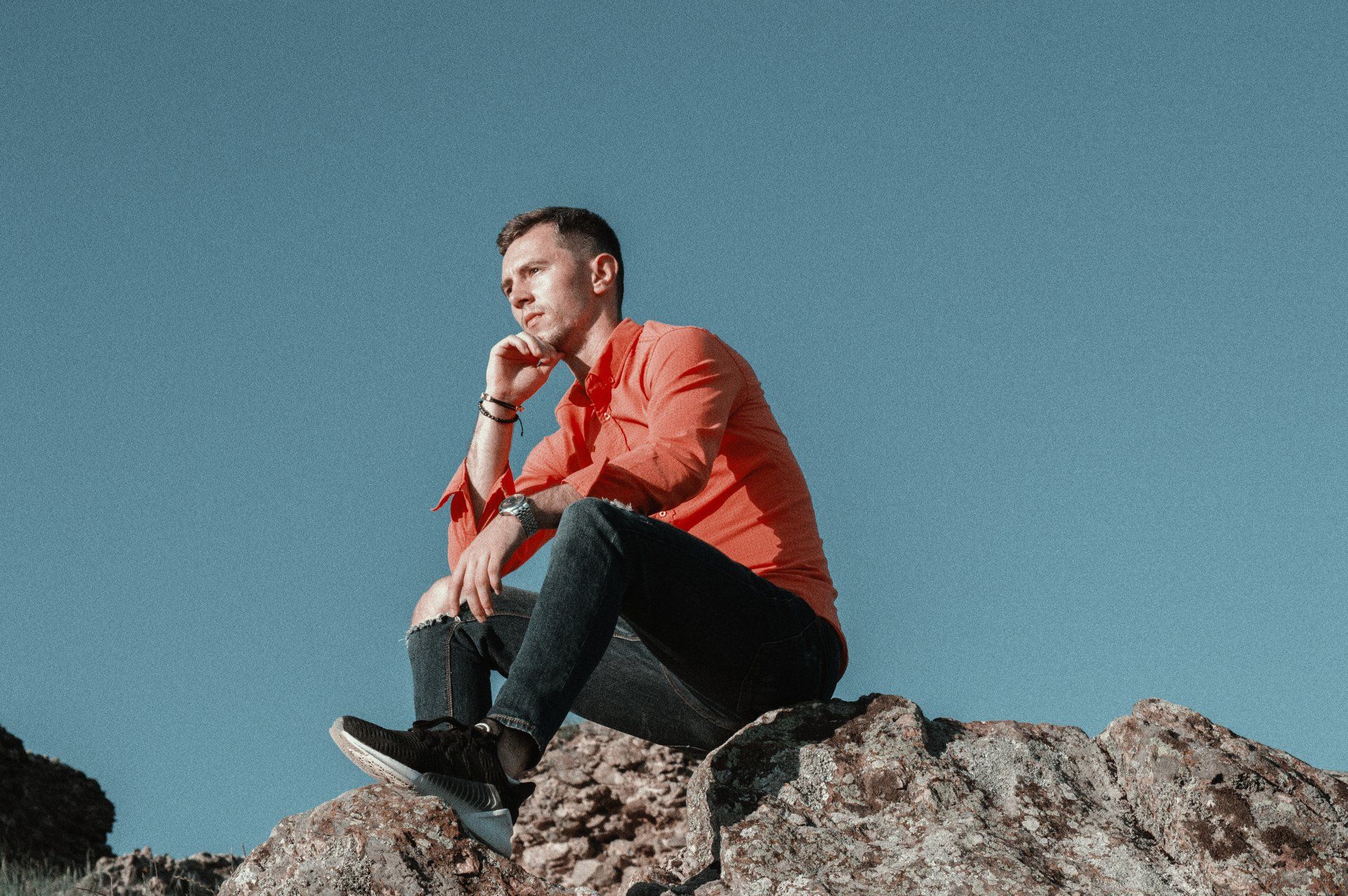 Man sitting on a rock, looking thoughtful, wearing an orange shirt and jeans. Blue sky background.