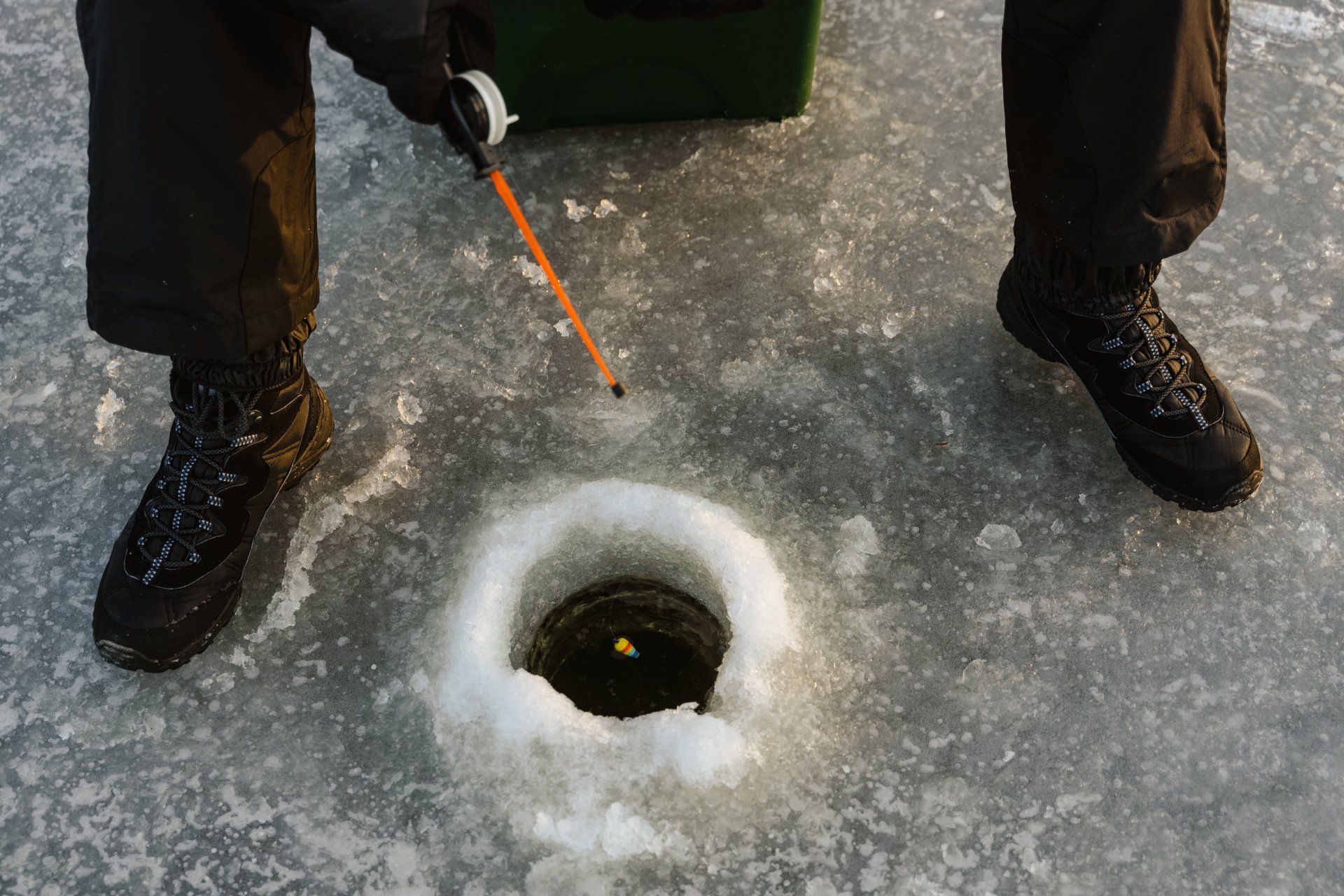 Person ice fishing: legs and boots surround a hole in ice, rod in hand, green tackle box nearby.