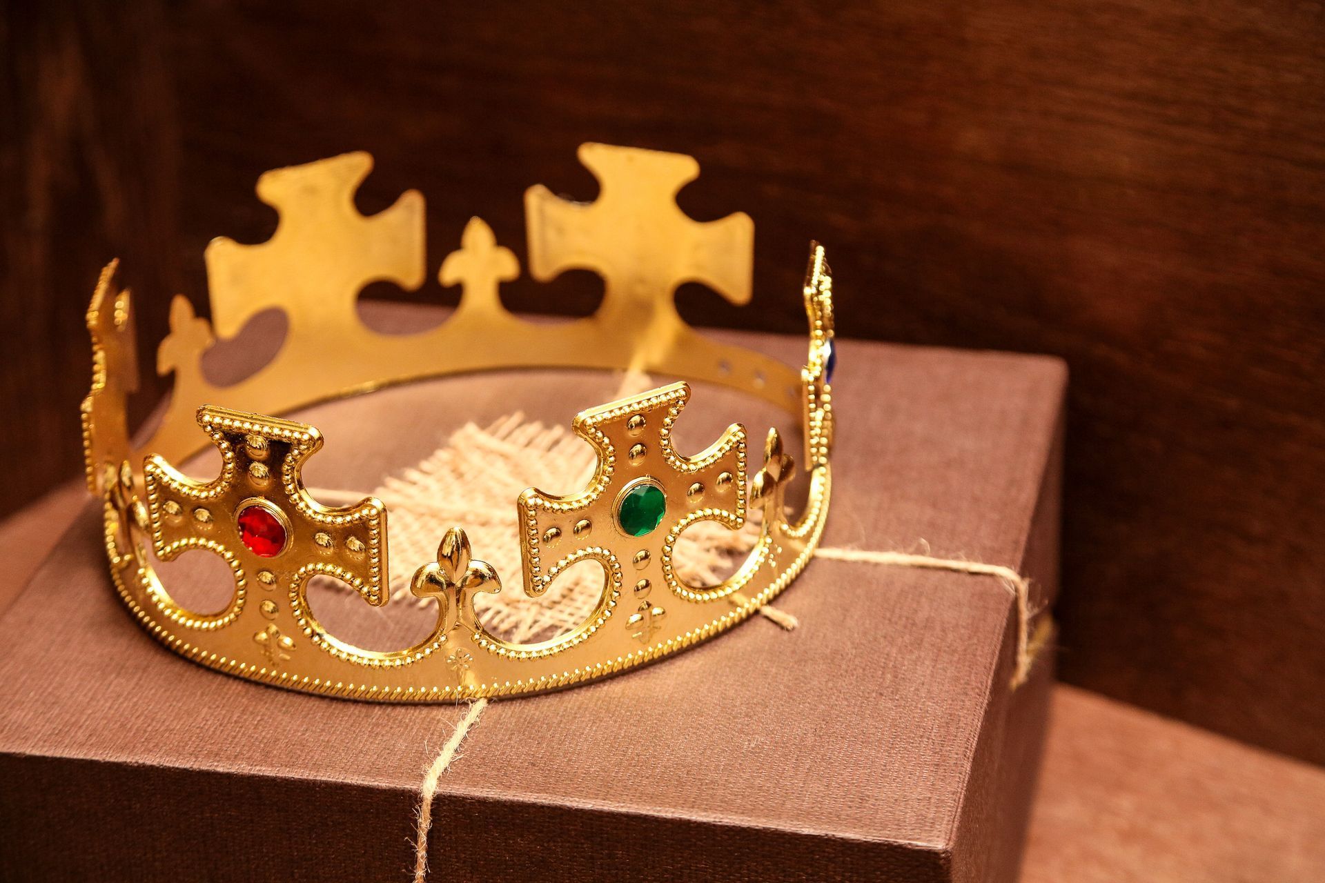 Gold crown with red and green gems on a brown box tied with twine, set against a wooden background.