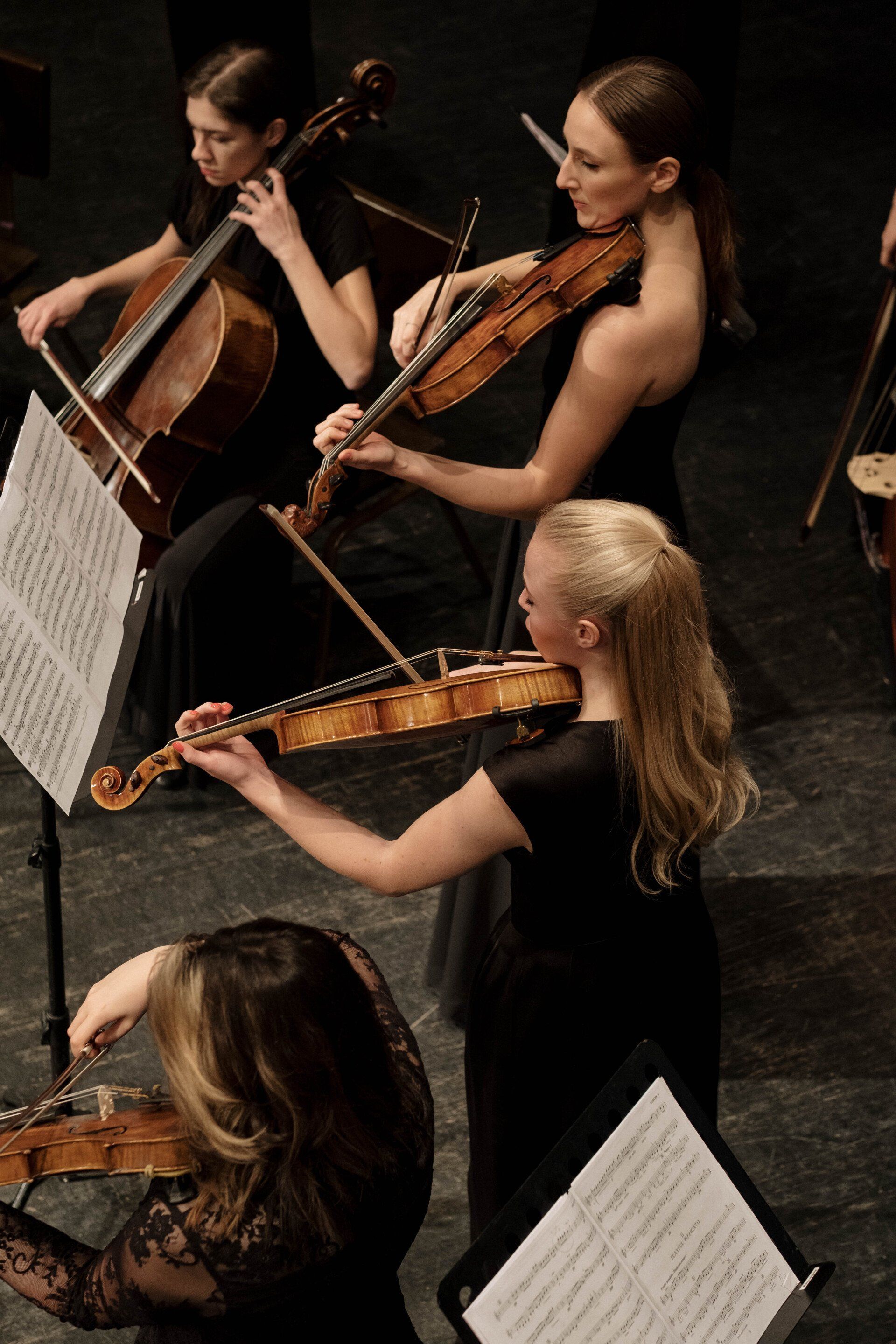 Orchestra members playing string instruments on stage, wearing black dresses.