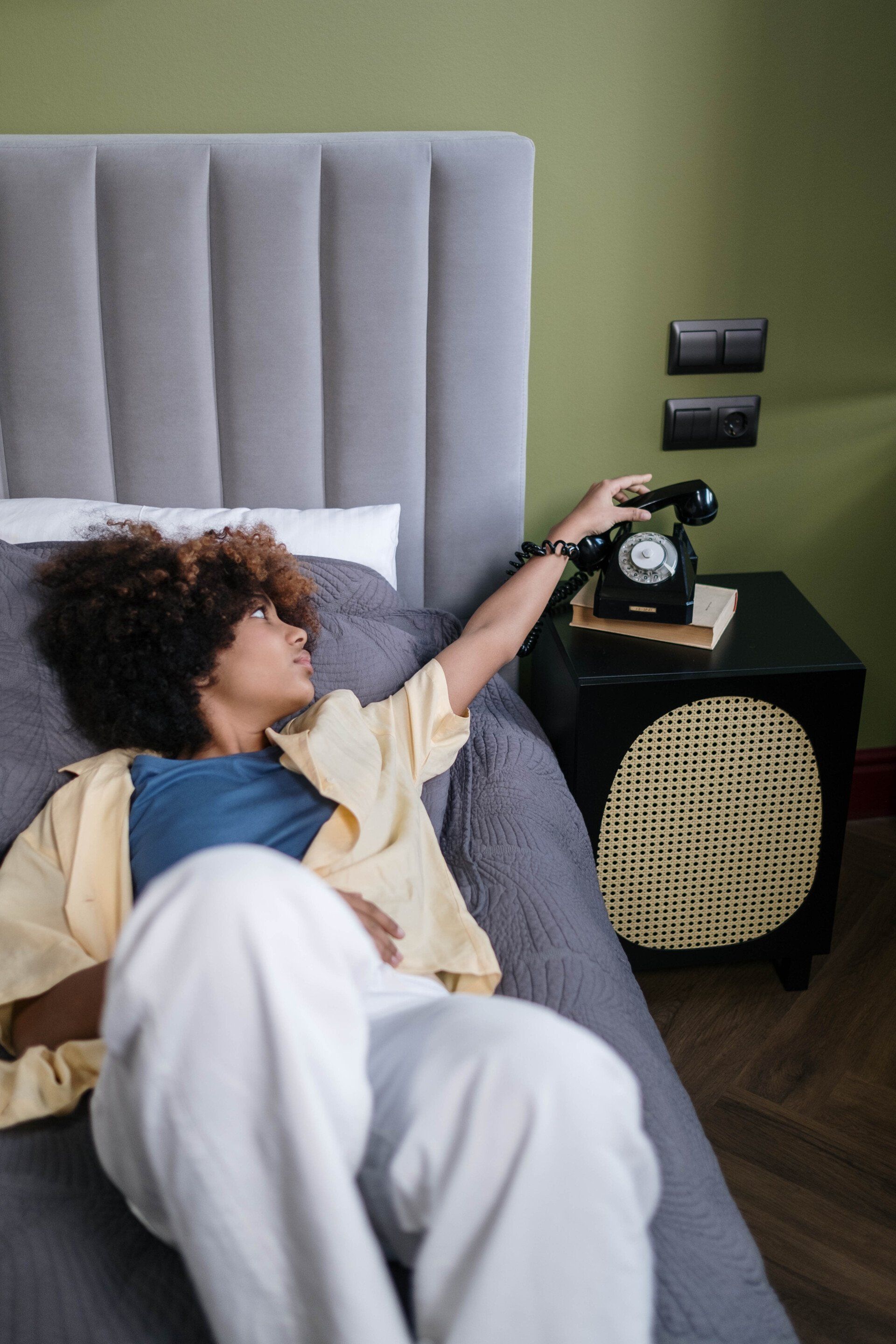 Person reaching for a black rotary phone on a nightstand beside a bed.