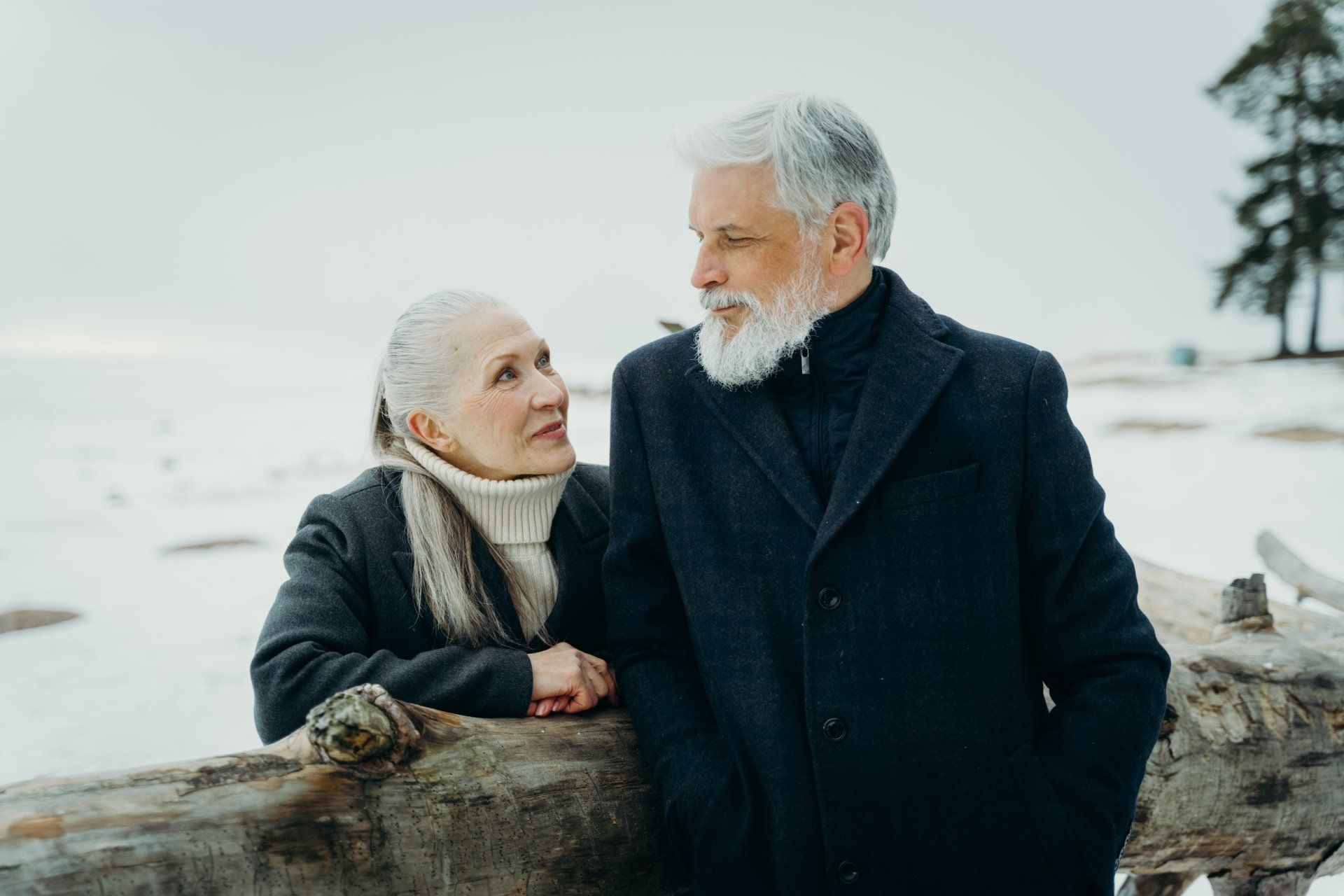 Elderly couple in winter coat looking at each other, leaning on a wooden beam, snow and trees in background.