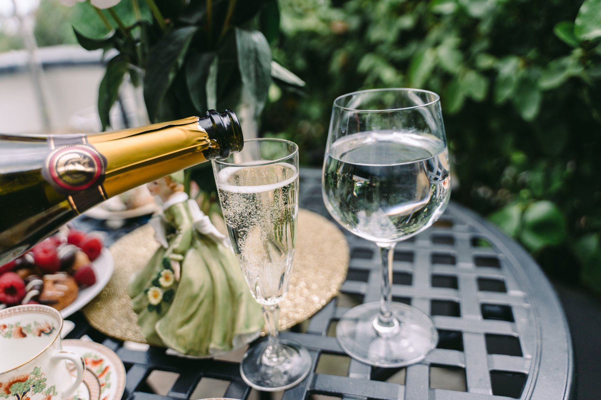 Champagne being poured into a glass, with another glass and pastries on a table outside.