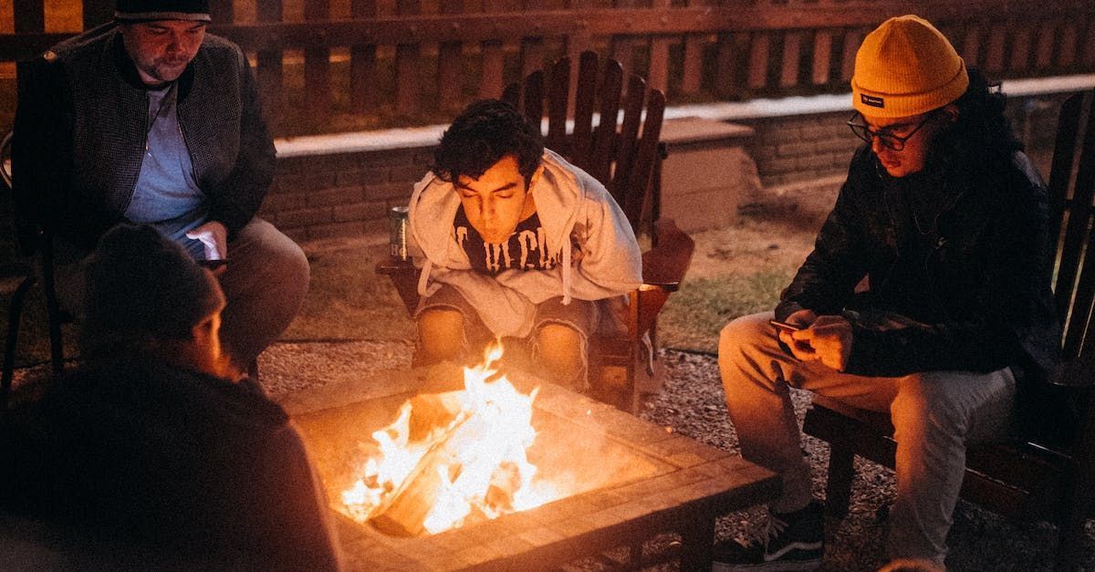 Group of people sit around a fire pit at night, looking intently at the flames.