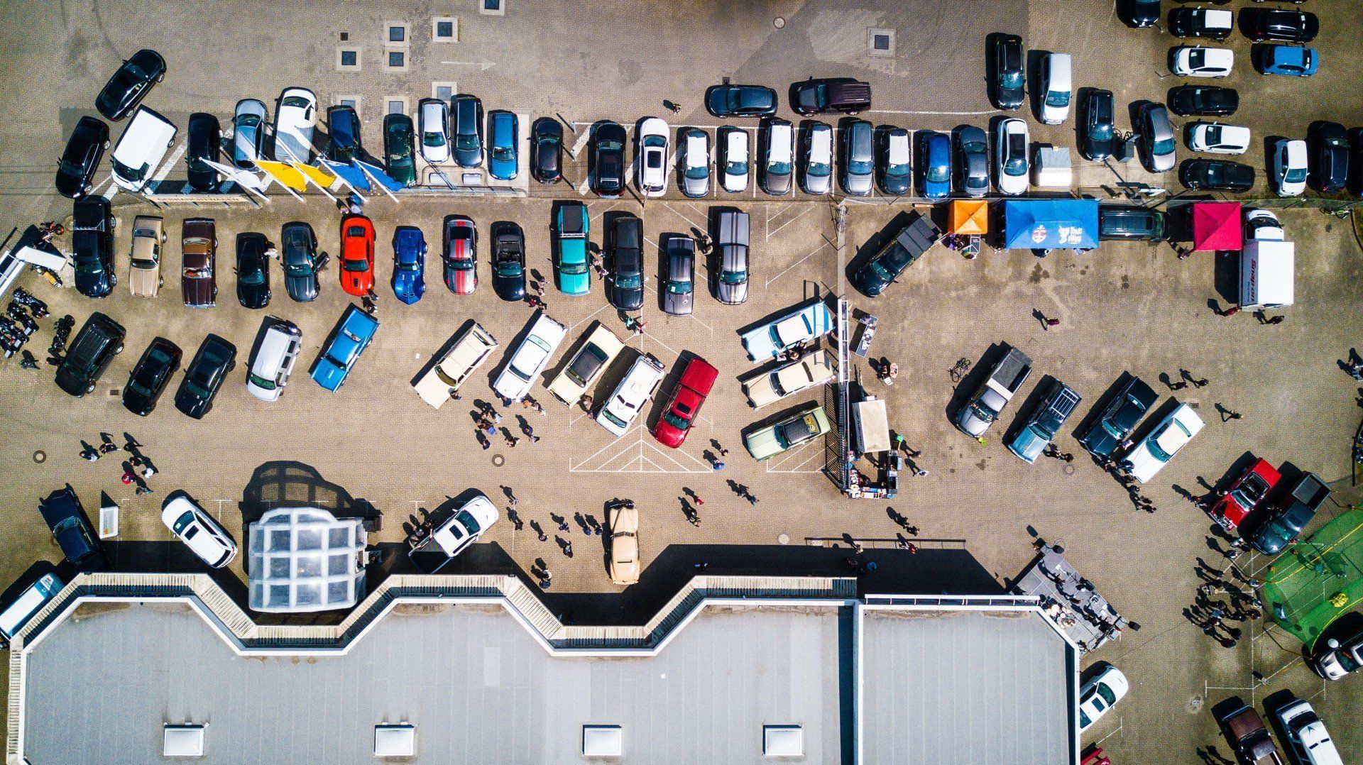 Aerial view of cars parked in an outdoor lot, next to a building. People stand around.