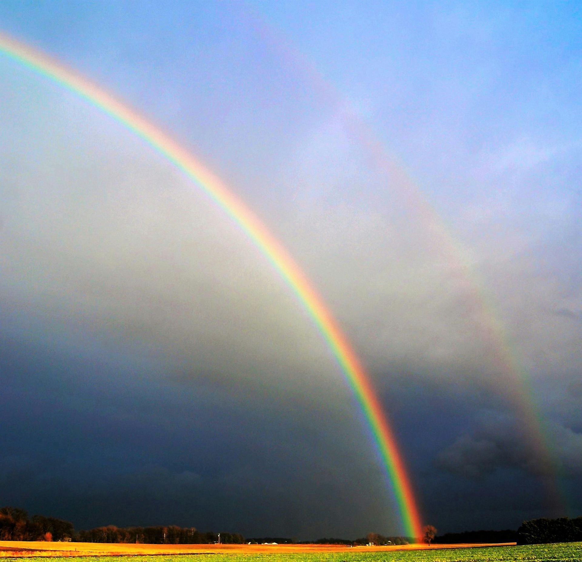 Double rainbow arcs across a dark, stormy sky over a field.