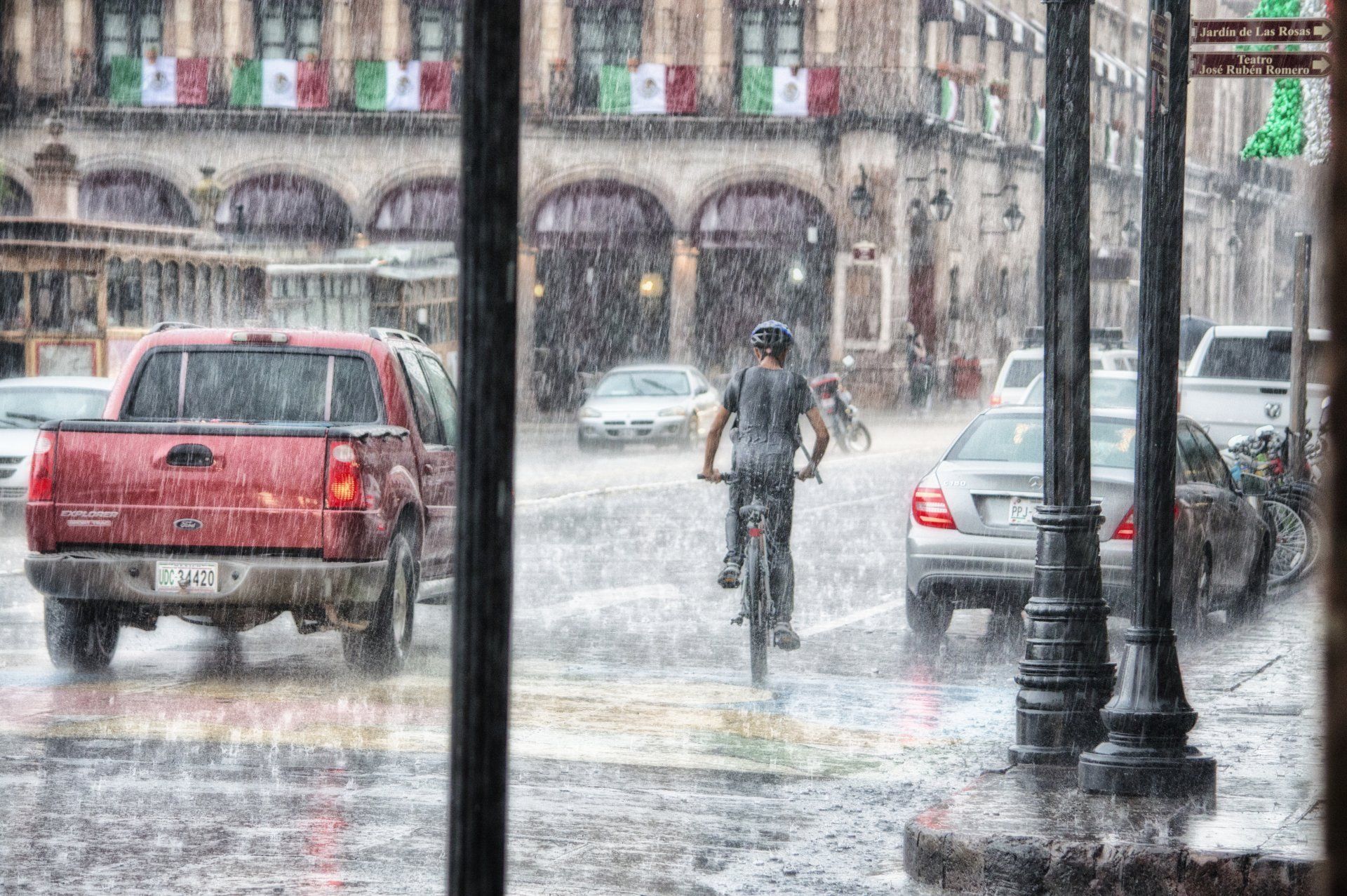 Rainy city street with a cyclist, cars, and buildings with Mexican flags.