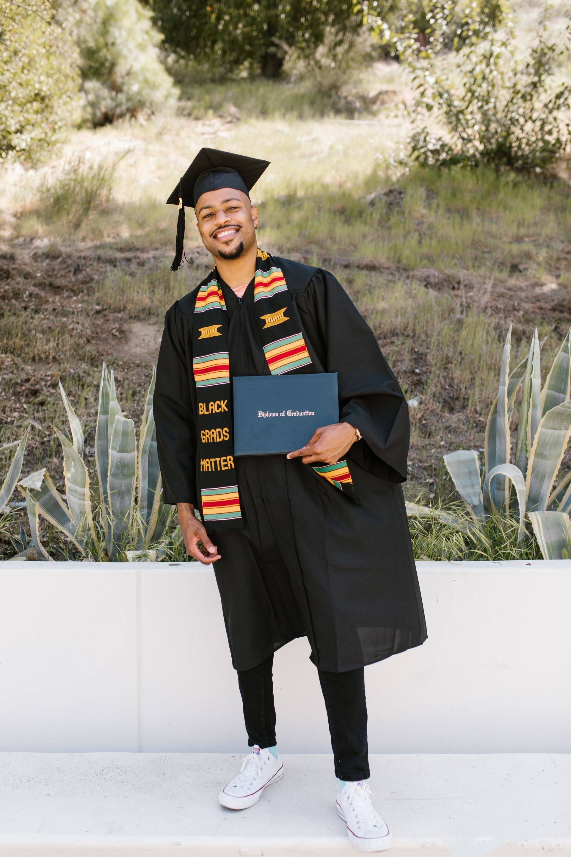Man in graduation cap and gown holding diploma, smiling outdoors.