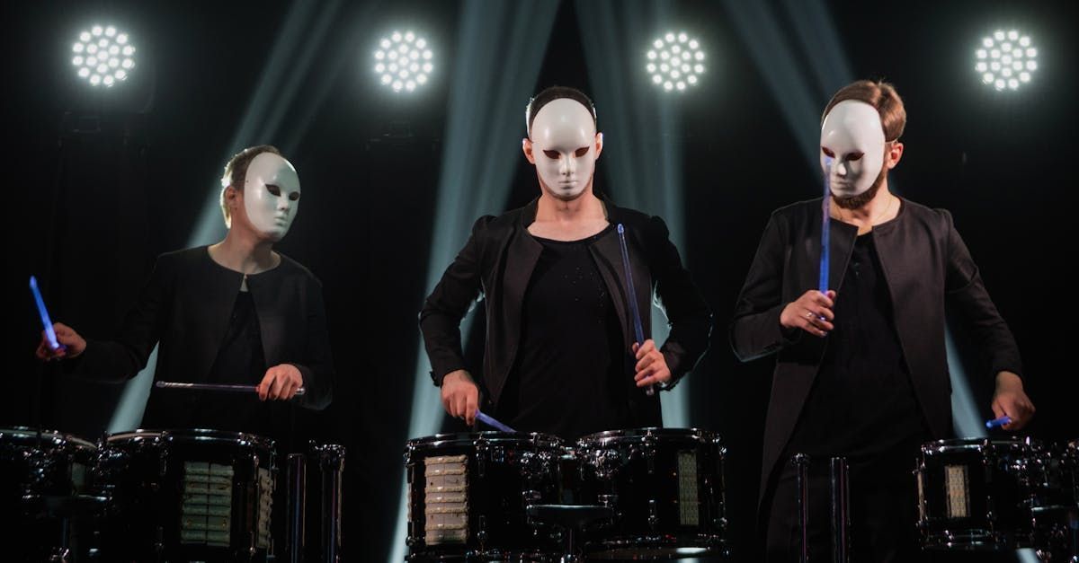 Three people wearing white masks playing drums with blue drumsticks on a stage.