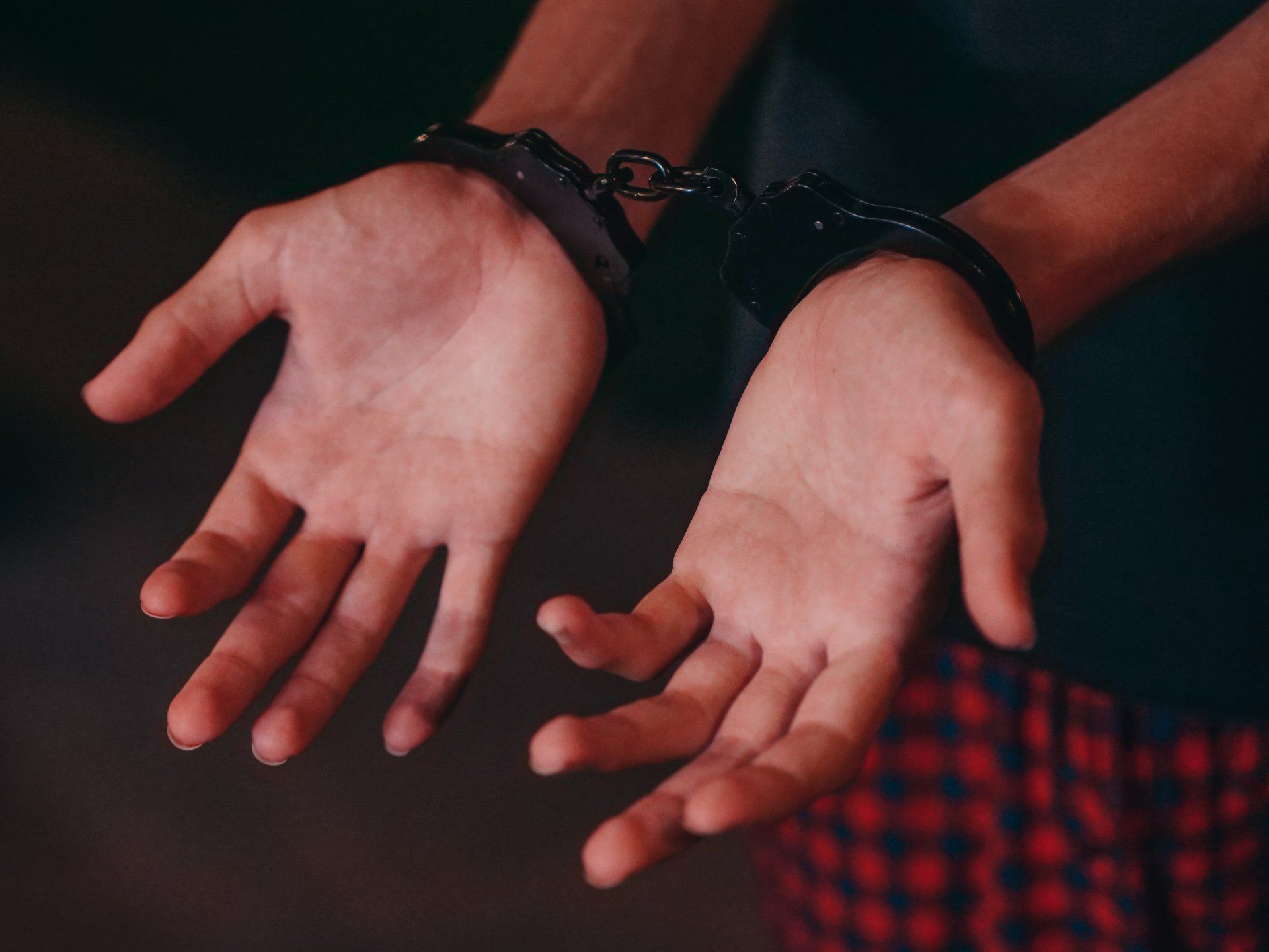 Person's hands in handcuffs, dark setting, wearing plaid pants and a dark shirt.