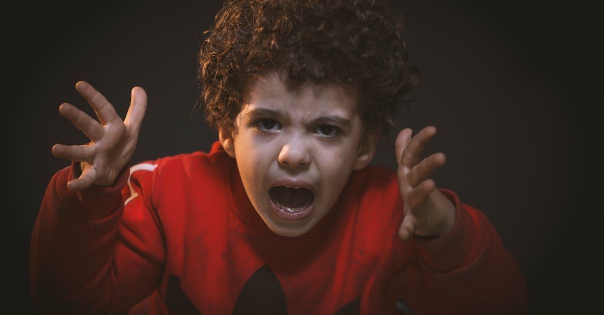 Child with curly hair yelling, arms raised, wearing a red sweatshirt against a dark background.