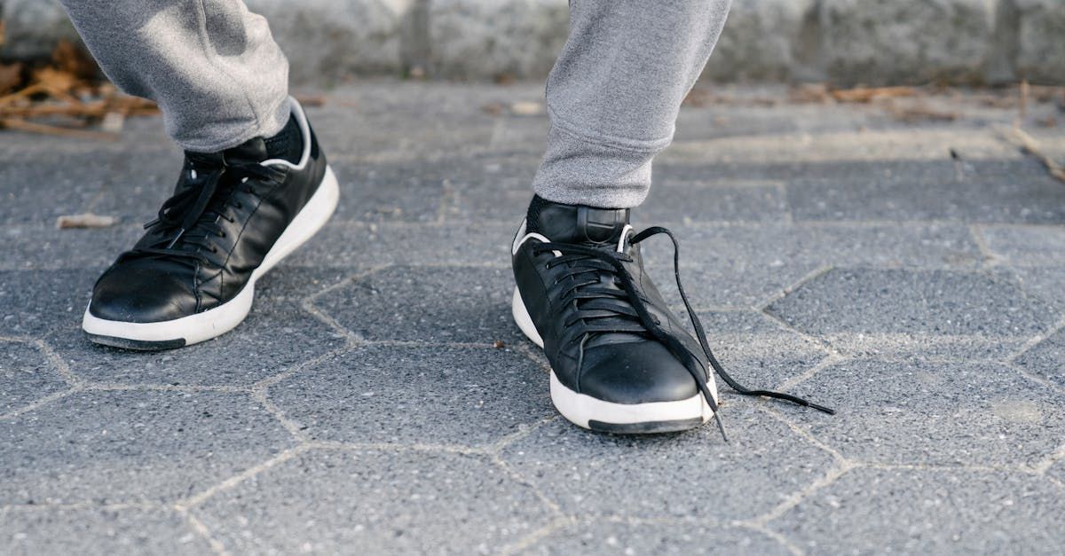 Person's feet with black sneakers, one shoelace untied, on a gray hexagonal paved surface.