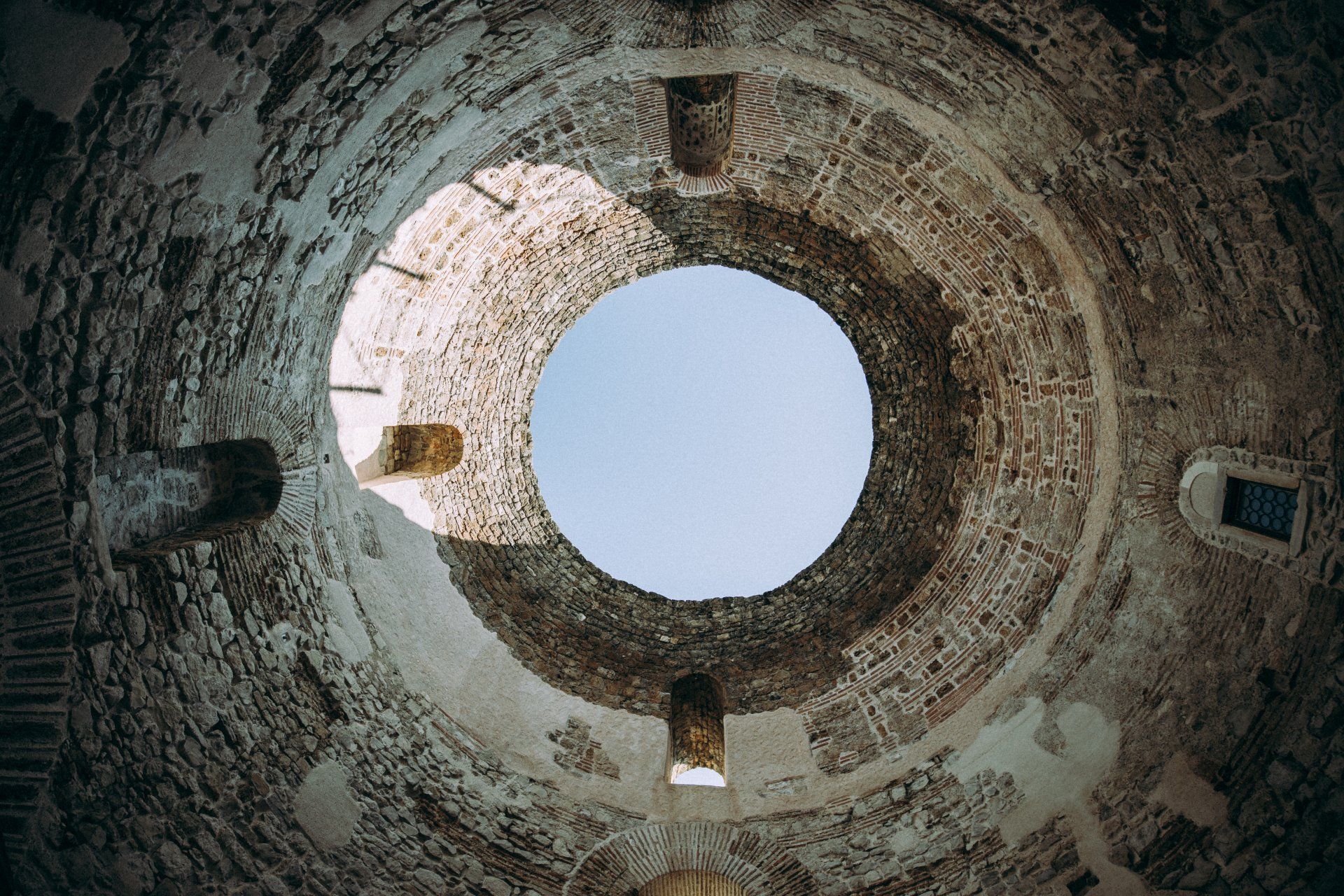 View looking up at a circular stone tower opening to blue sky. Sunlight illuminates the interior.