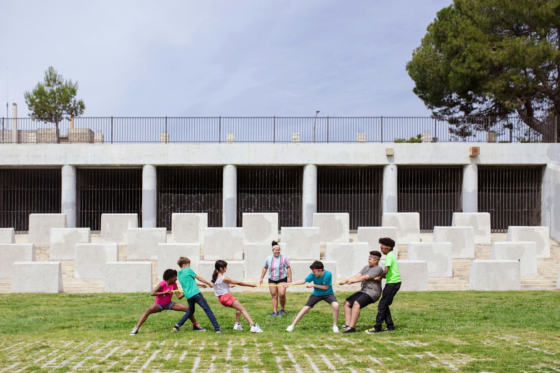 Children playing tug-of-war on grass, concrete blocks in the background. Sunny day.