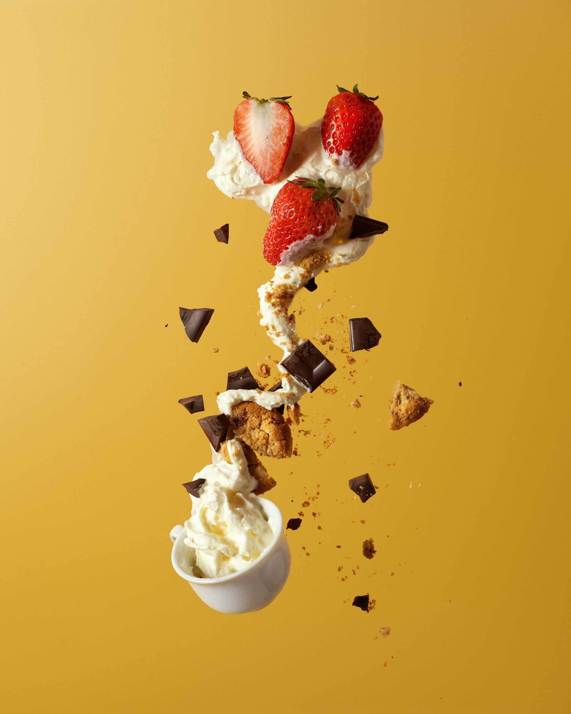 Whipped cream, strawberries, chocolate, and crumbs appear suspended in mid-air against a yellow backdrop.
