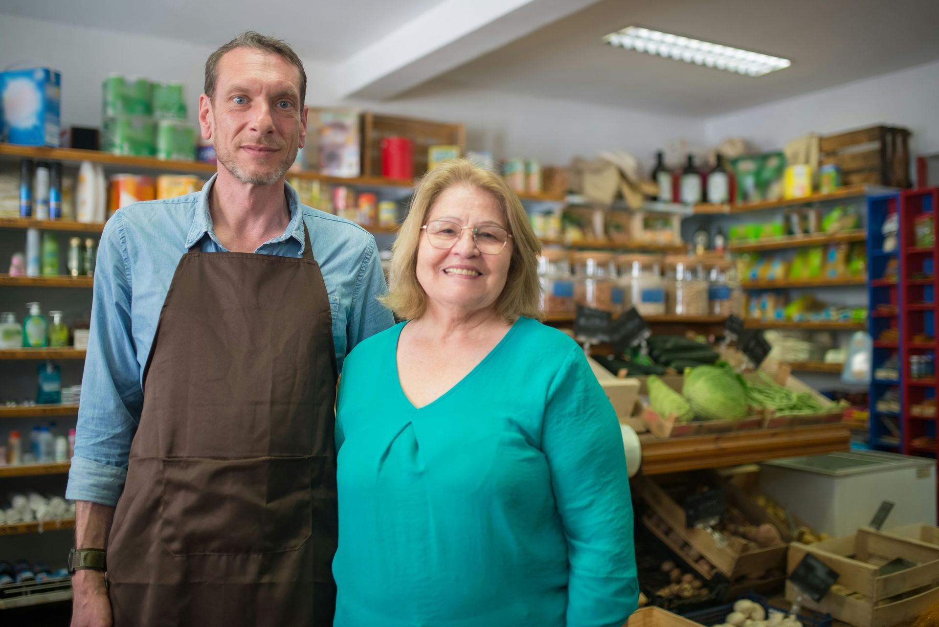 Two people stand in a small grocery store. Man in apron, woman in teal shirt. Shelves stocked with products.