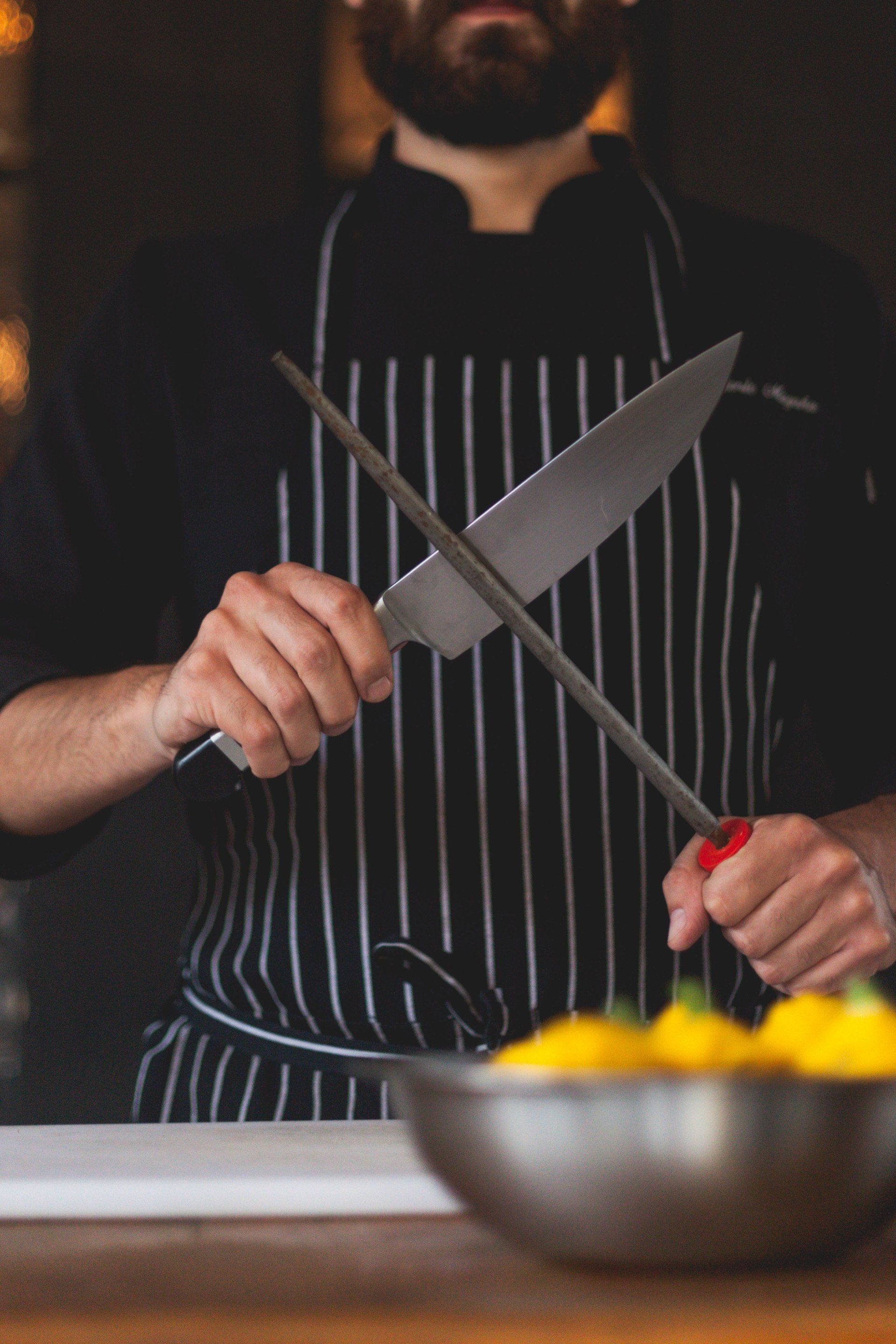 Chef sharpening a knife with a steel rod, wearing a striped apron, over a bowl.