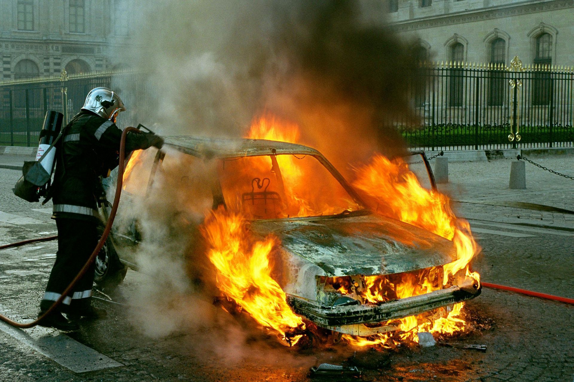 Firefighter extinguishing a car engulfed in flames with smoke billowing.