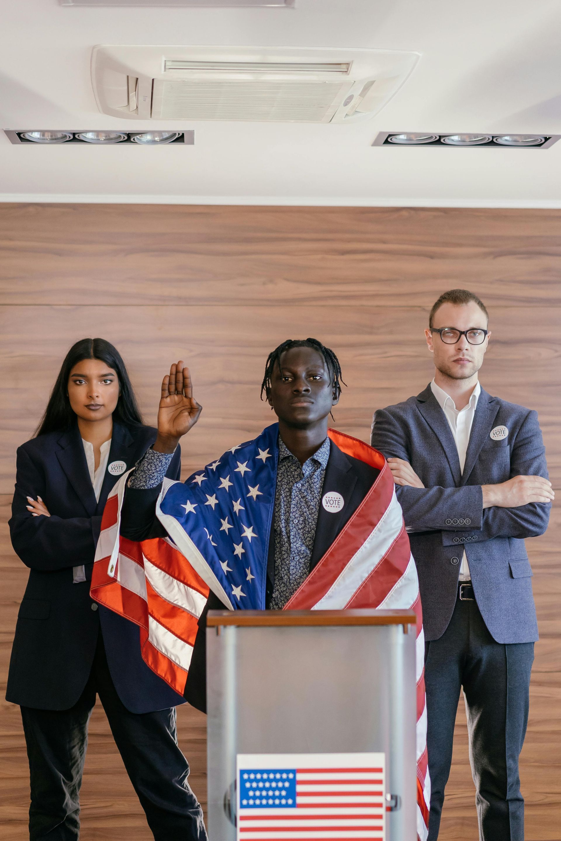 Three people in suits; one draped in a US flag, speaking at a podium, the others standing on either side.