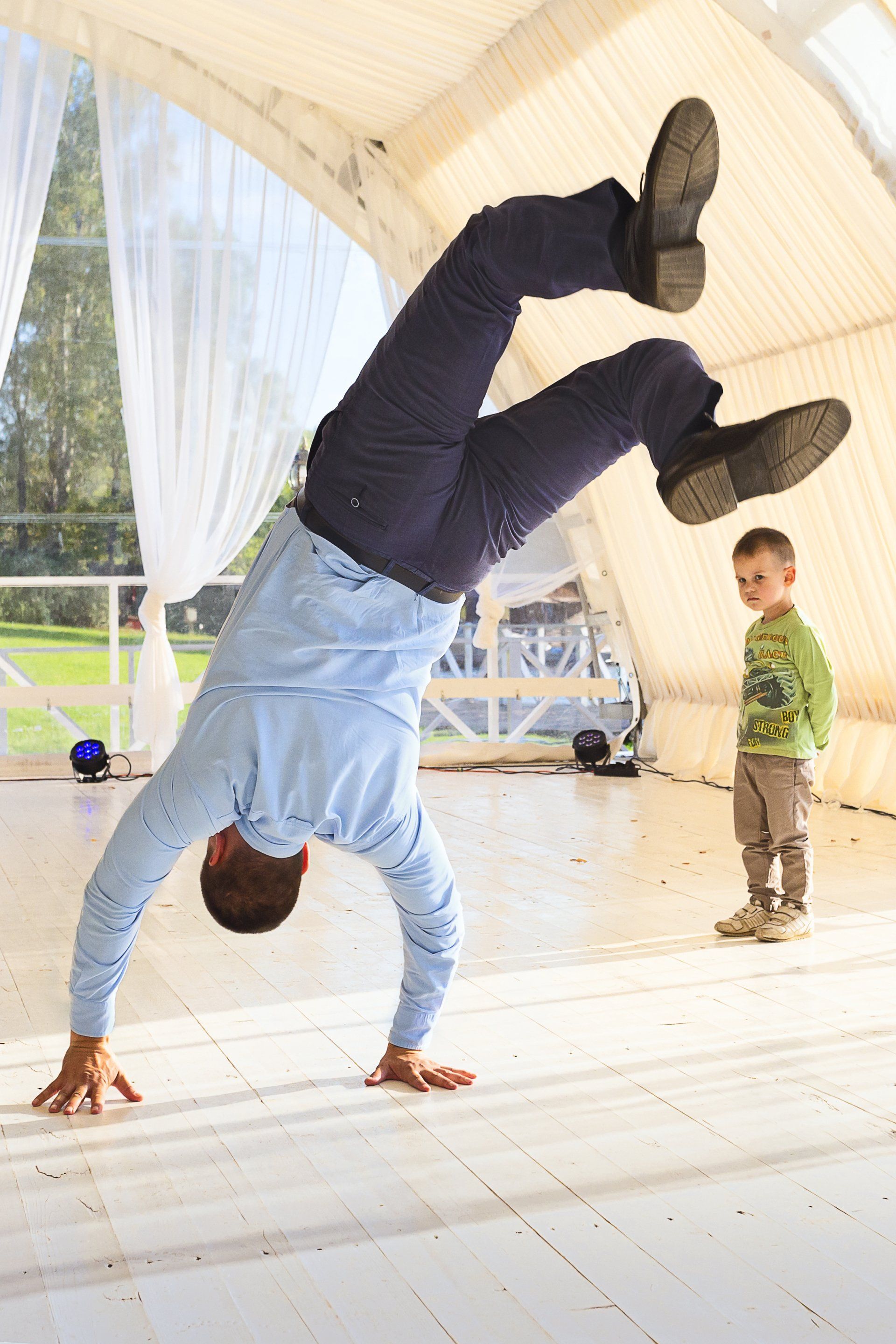 Man doing a handstand, feet in the air, indoors; child watches.