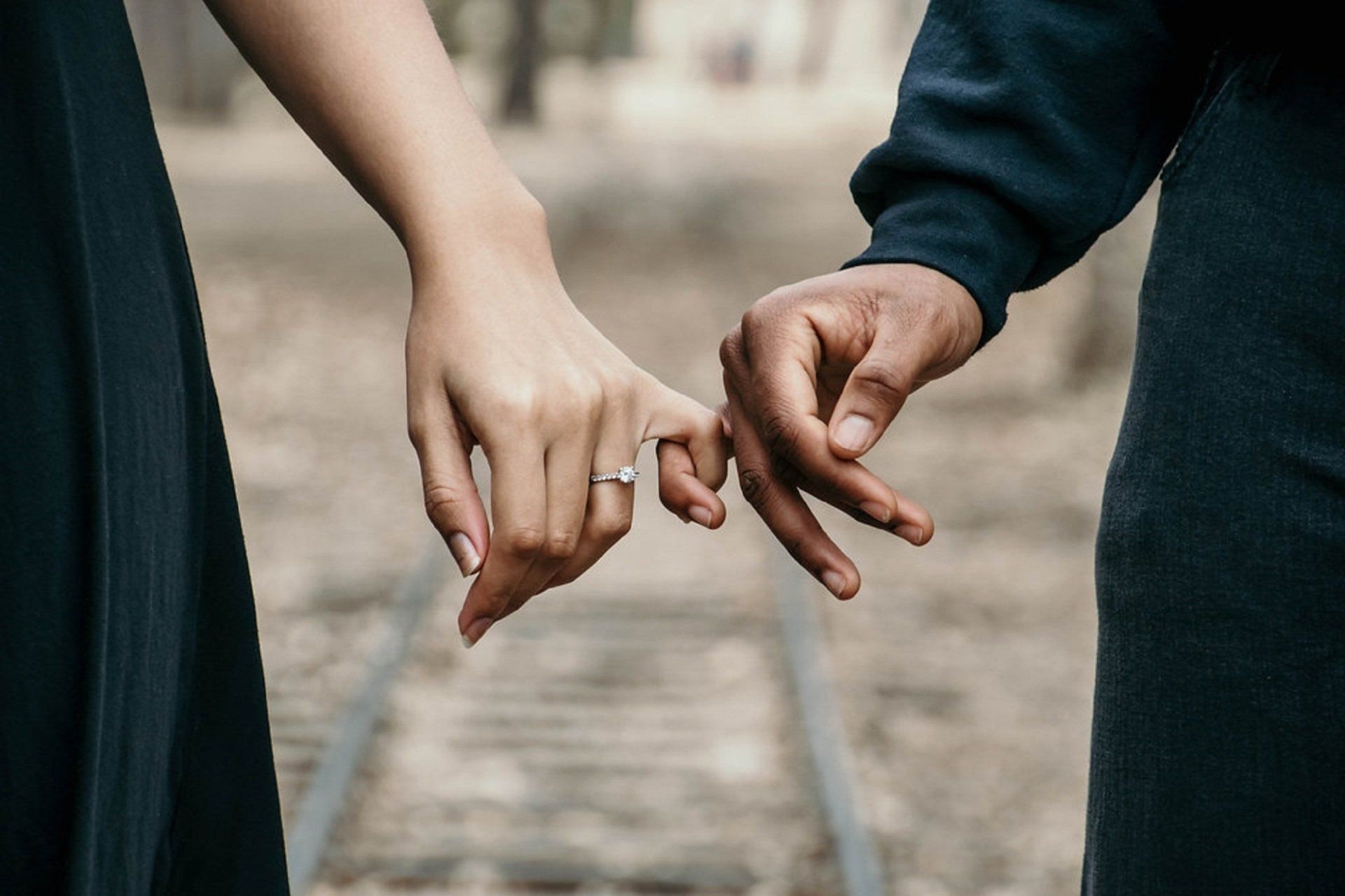 Two people holding hands, pinky fingers interlocked. One wears a ring. Blurred outdoor background.