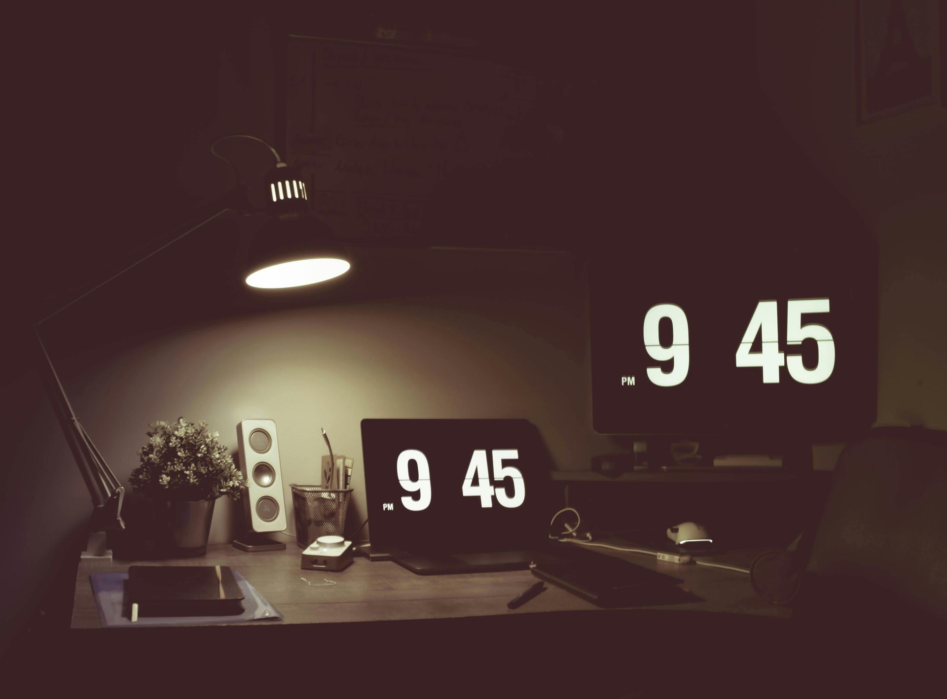 Desk with digital clocks displaying 9:45, a lamp, speakers, and a plant. Dark setting.
