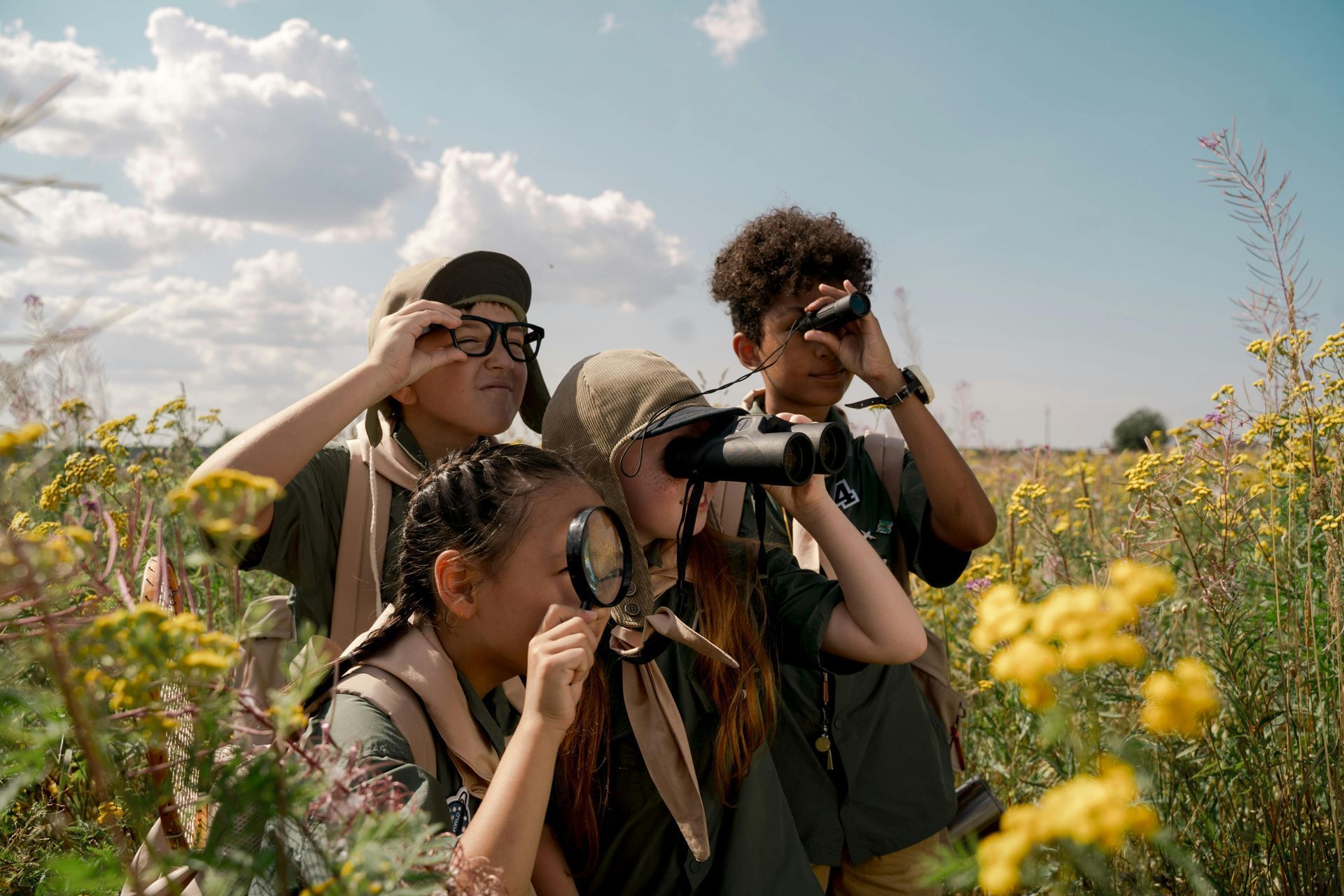Four children in field, using binoculars and magnifying glass, observing nature.