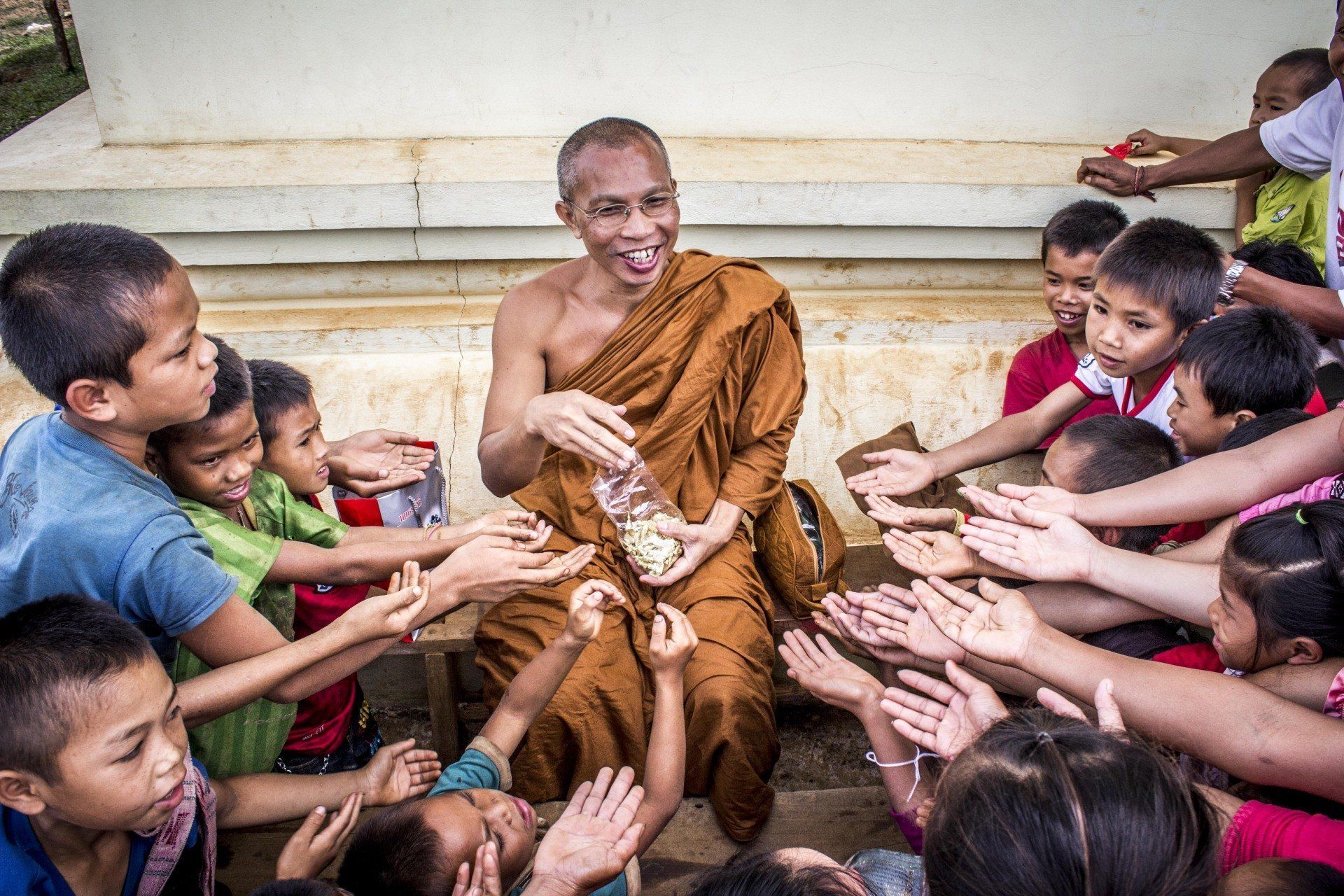 Monk in orange robes distributing food to a group of children with outstretched hands outdoors.