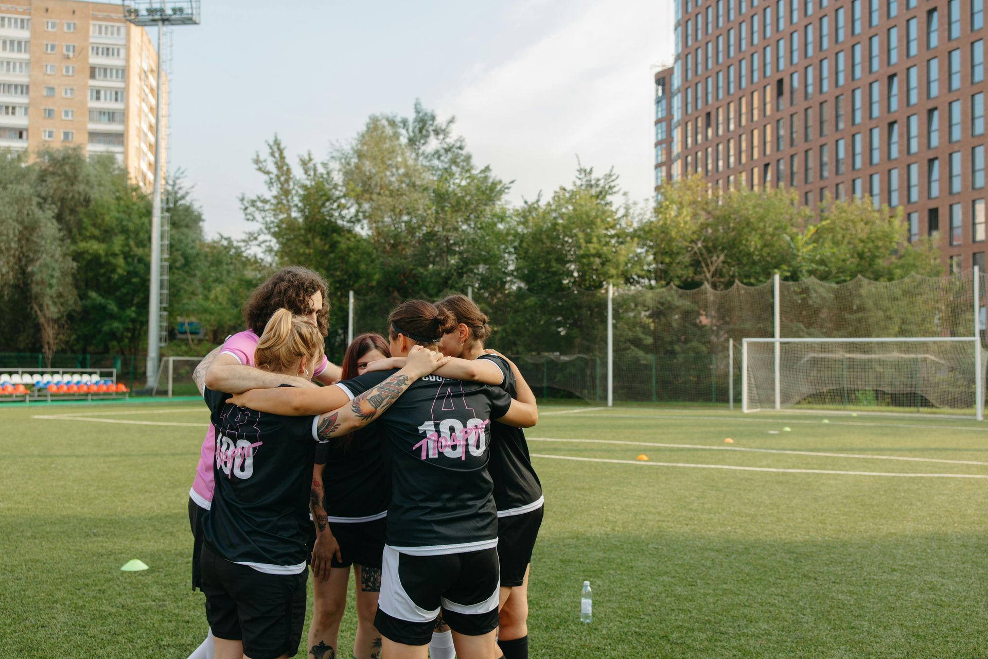 Group of athletes huddle on a green soccer field, wearing black jerseys and hugging, in front of buildings.