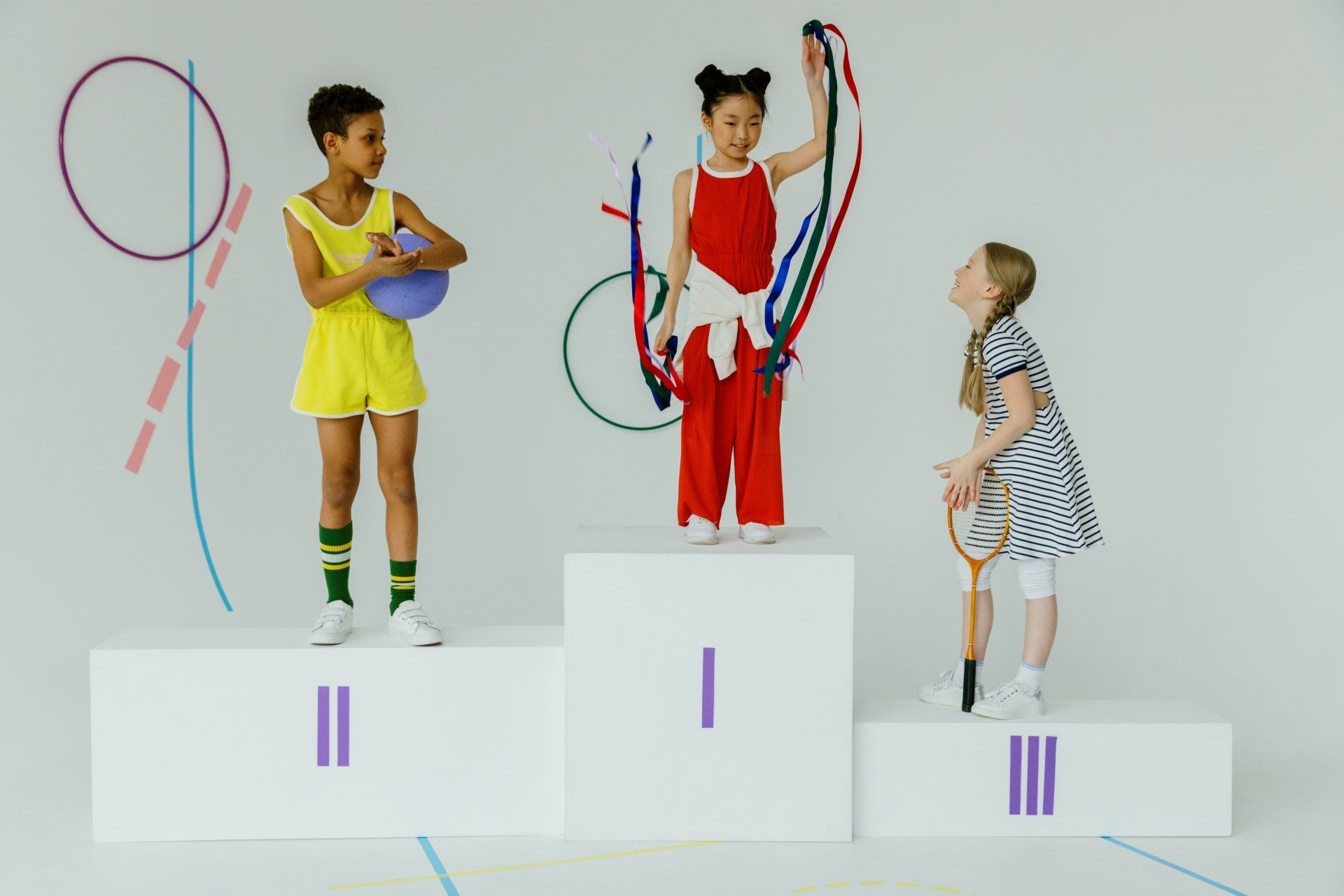 Children on podium; girl in red holding ribbons, others with ball and racket. Colored shapes on wall.