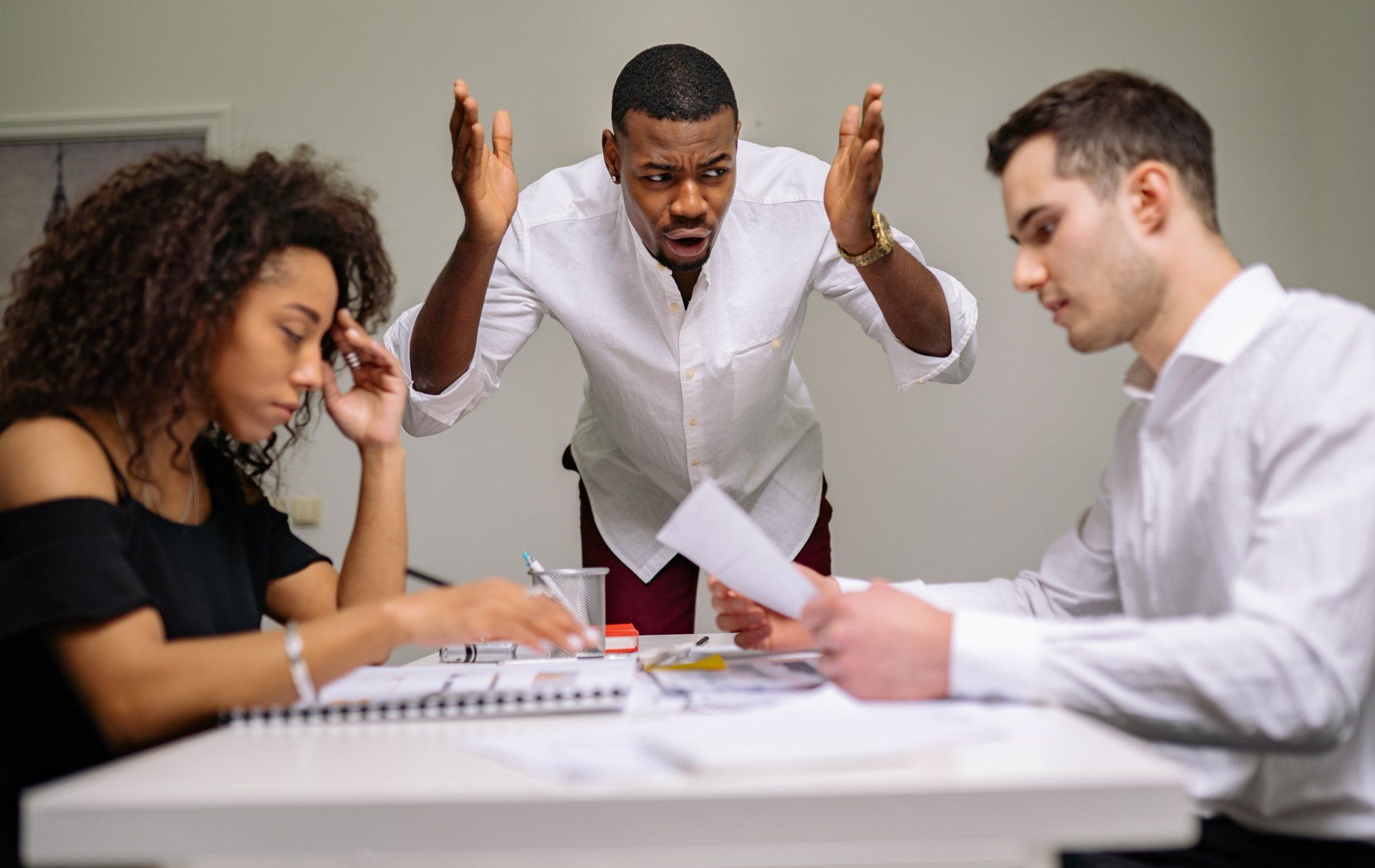 Three people in a tense meeting; man standing angrily, others looking stressed, documents on desk.