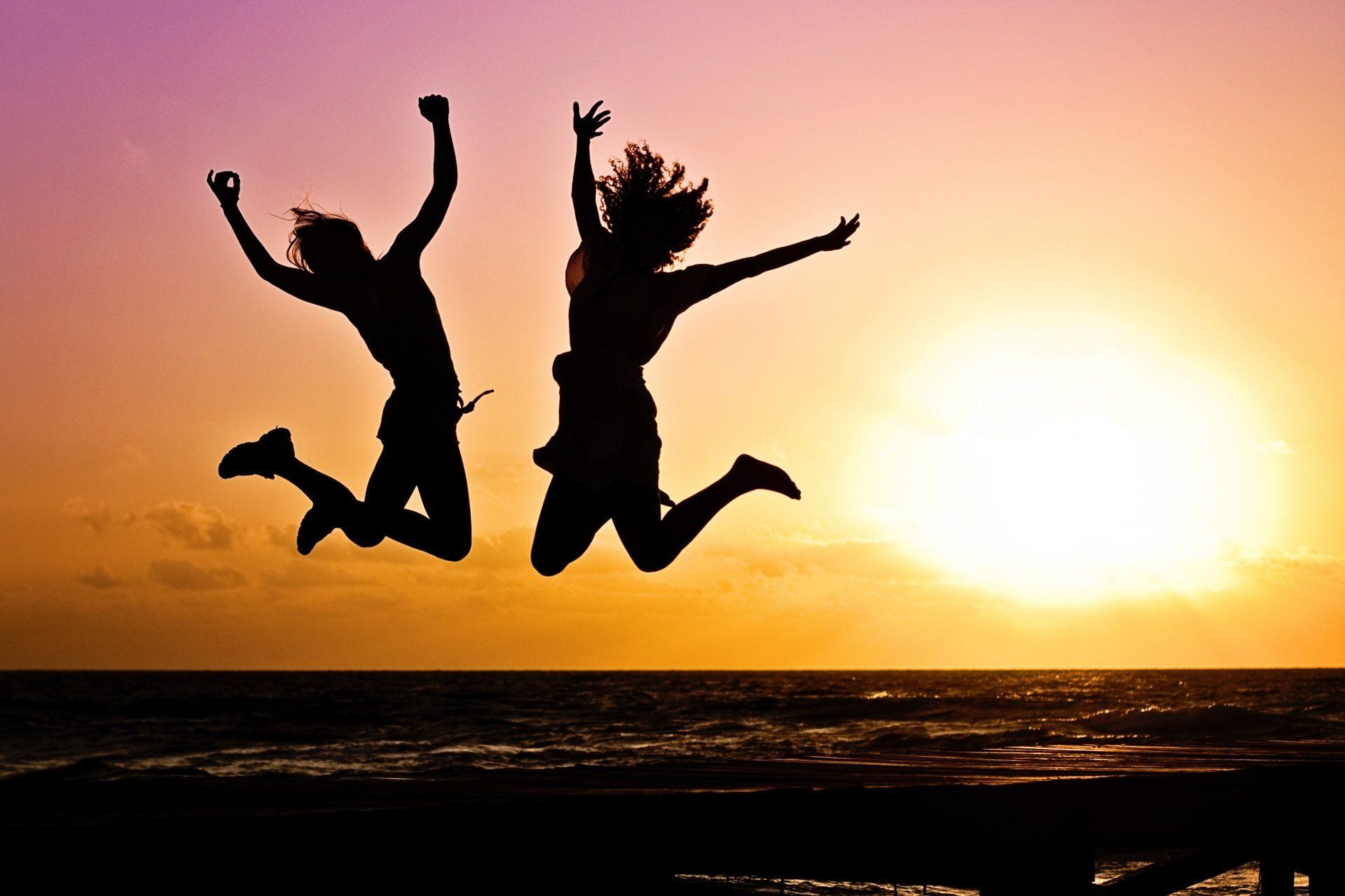 Two people silhouetted jumping for joy against a colorful sunset over the ocean.